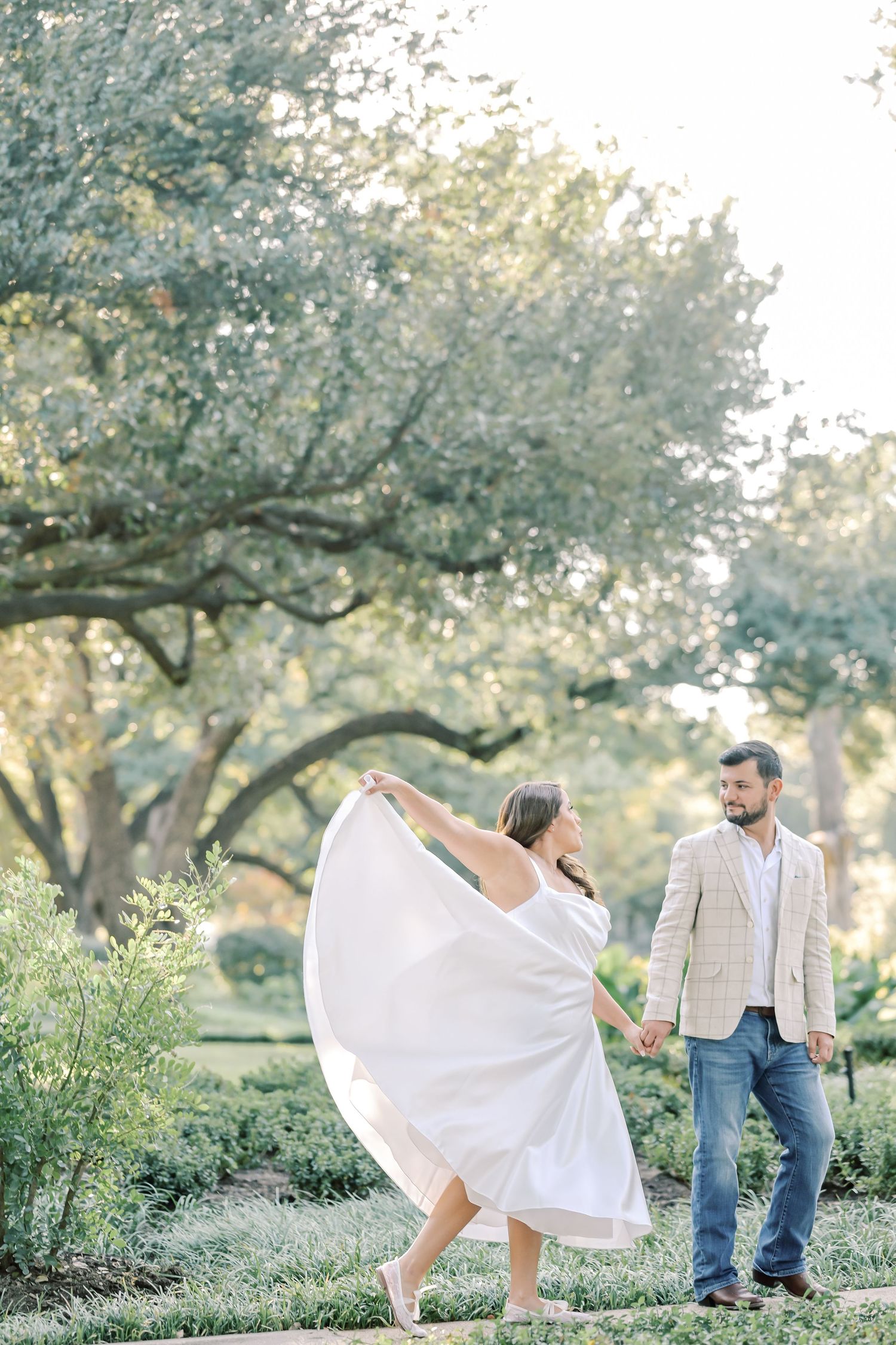 Woman in a flowing white dress twirls while holding hands with a smiling man in a plaid blazer during an outdoor engagement photo shoot.