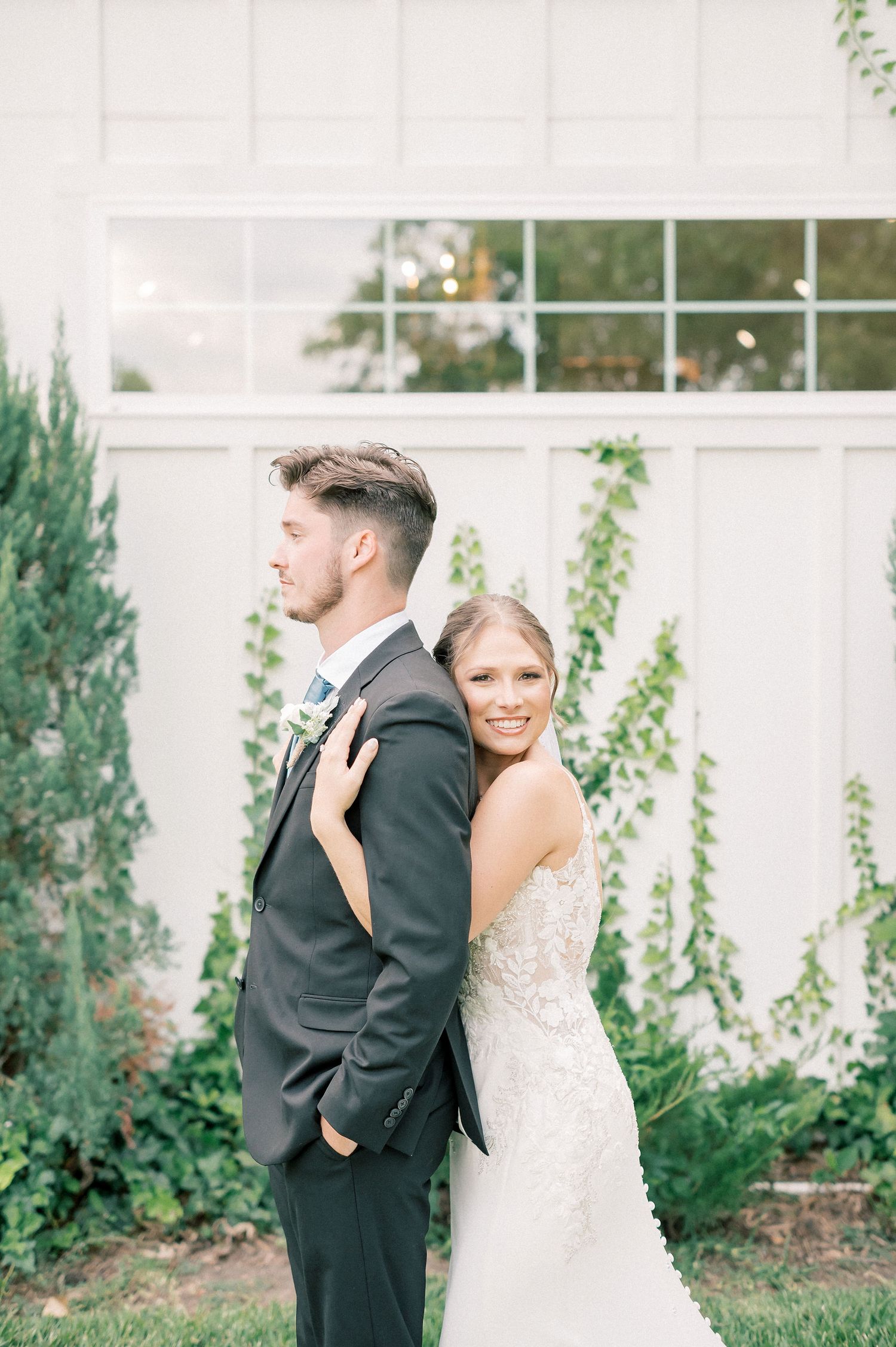 Romantic wedding portraits captured against a white exterior wall with lush green landscaping.
