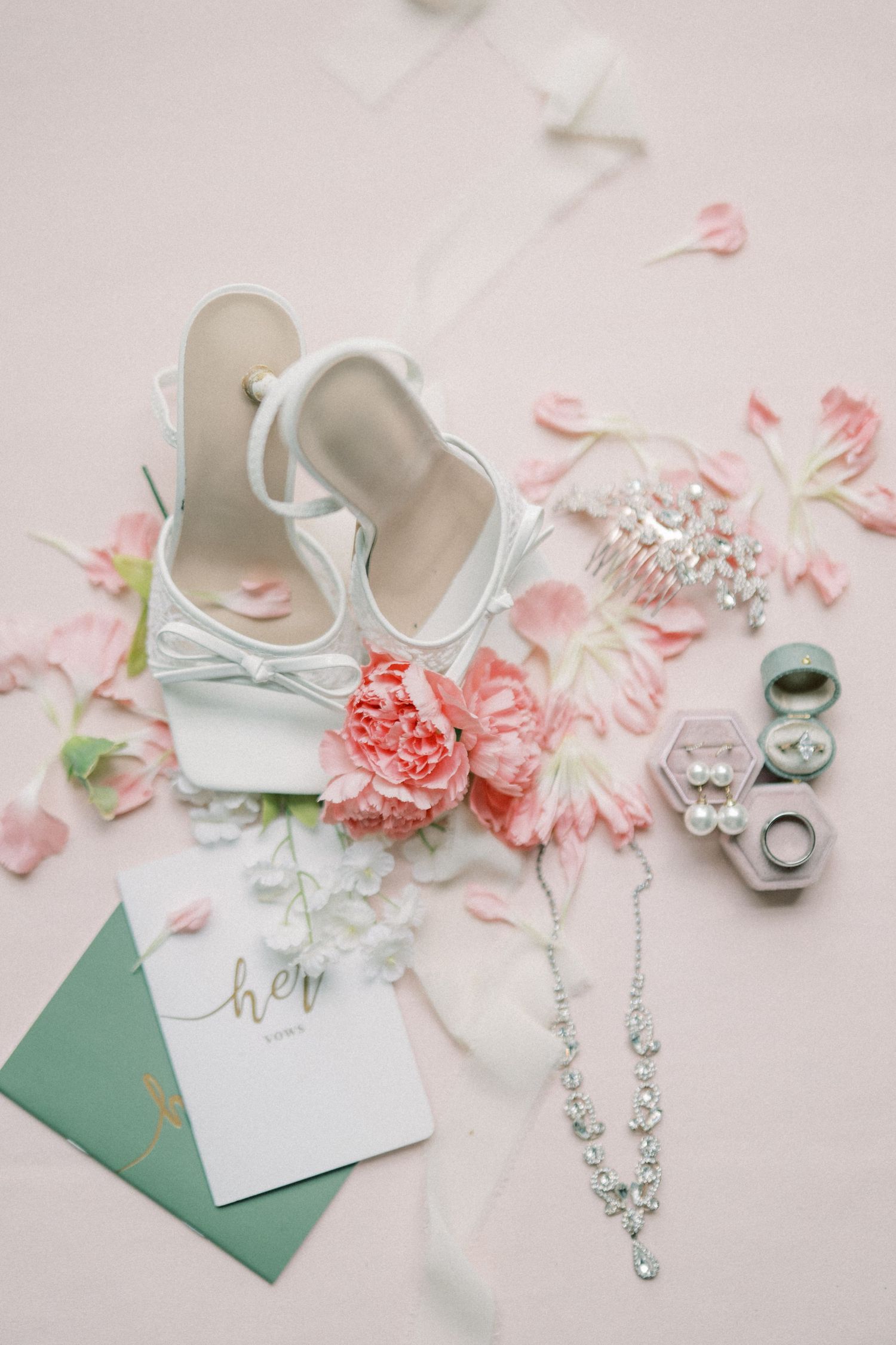 Elegant bridal flat lay with white heels, pink peonies, diamond necklace, earrings, ring boxes, and a green envelope on blush background.