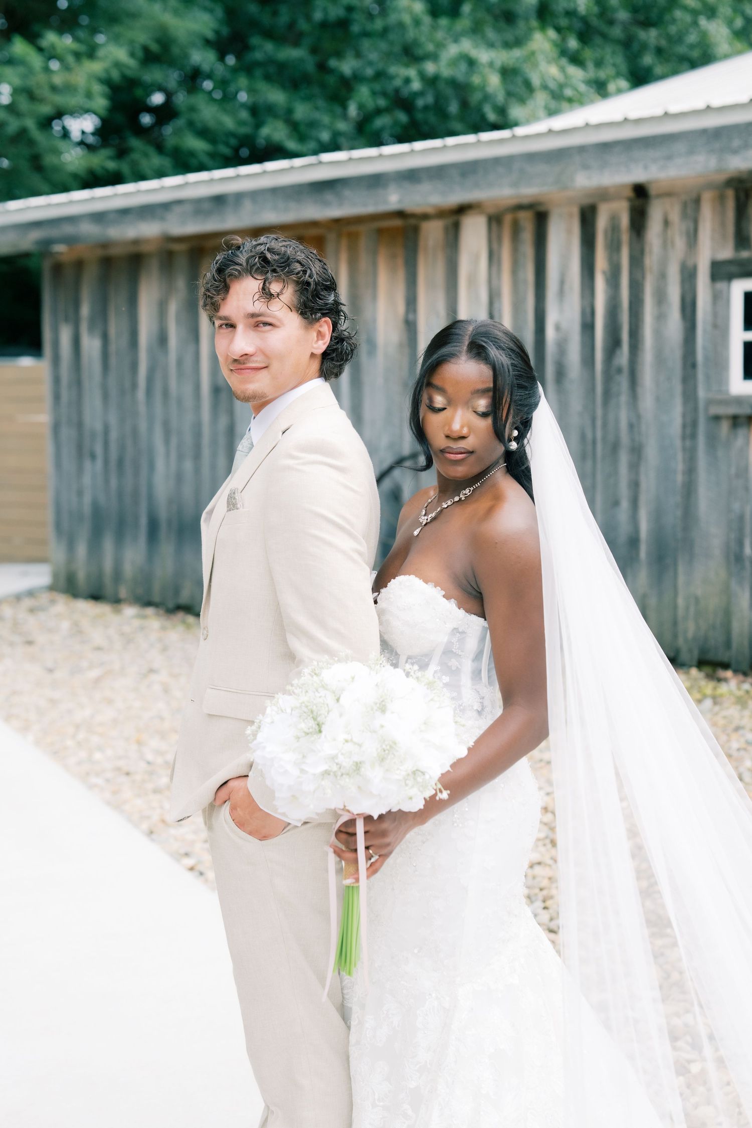 Interracial couple in wedding attire pose back-to-back, bride holding white bouquet with veil, groom in beige suit.