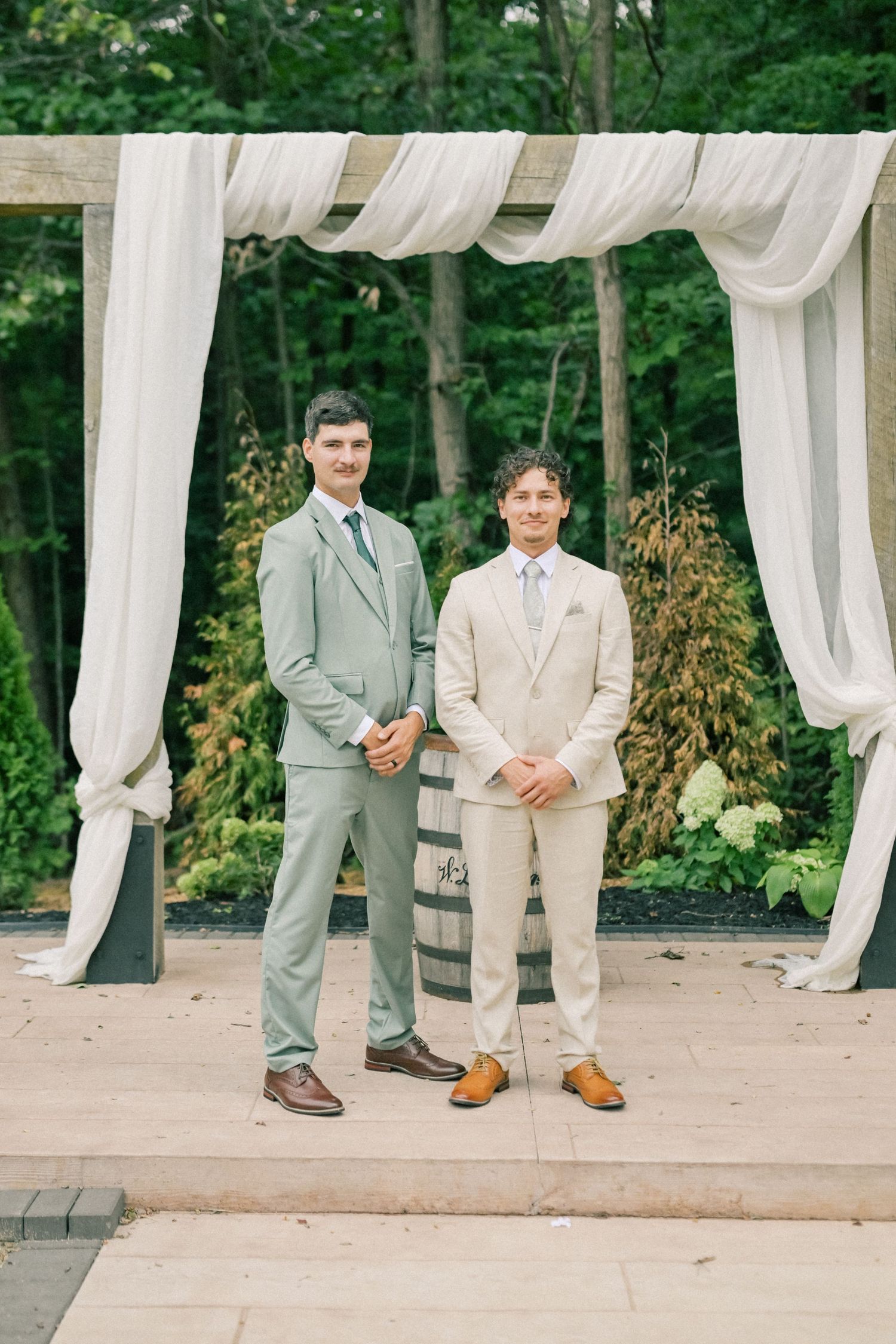 Two grooms in sage green and beige suits stand under a draped wooden arch at an outdoor wedding ceremony.