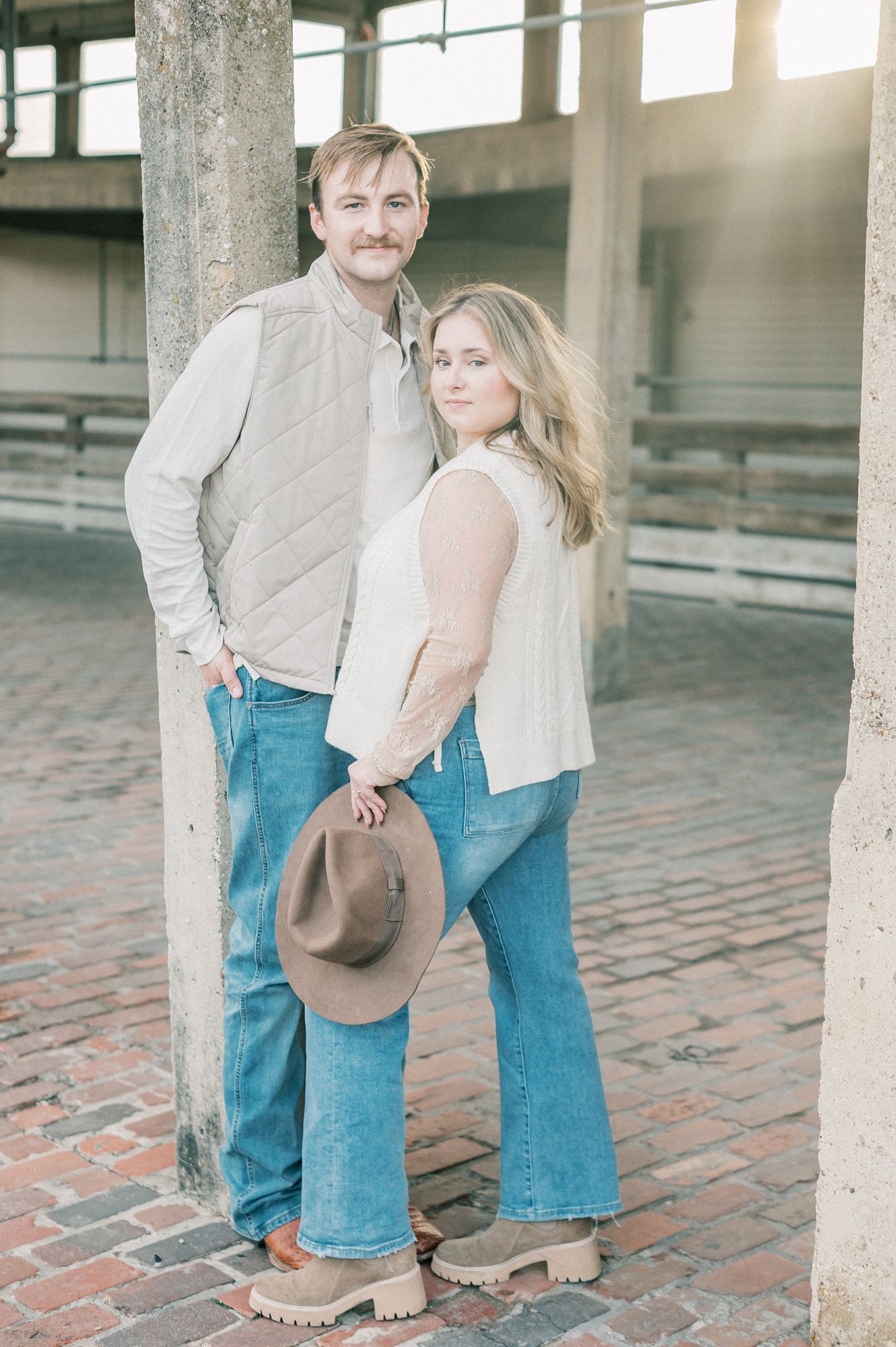 A couple in matching denim and beige outfits poses together on a brick patio near concrete pillars.