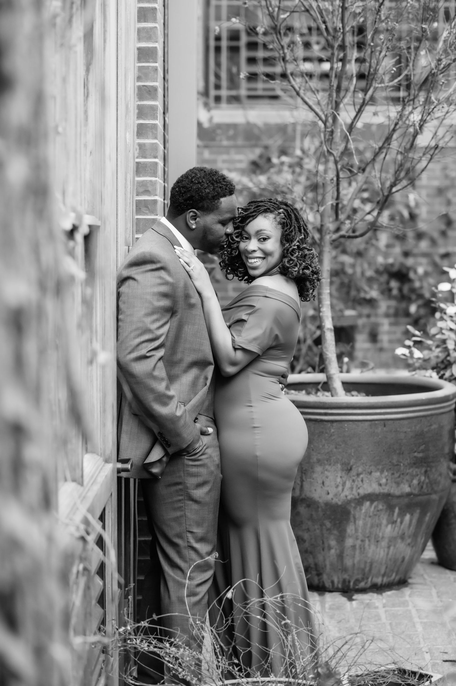 Black and white photo of a smiling couple in formal attire leaning against a wall in an outdoor garden.