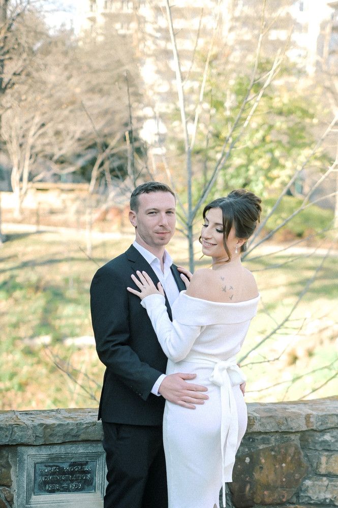 Bride in off-shoulder white gown embraces groom in black suit outdoors near stone wall in park.