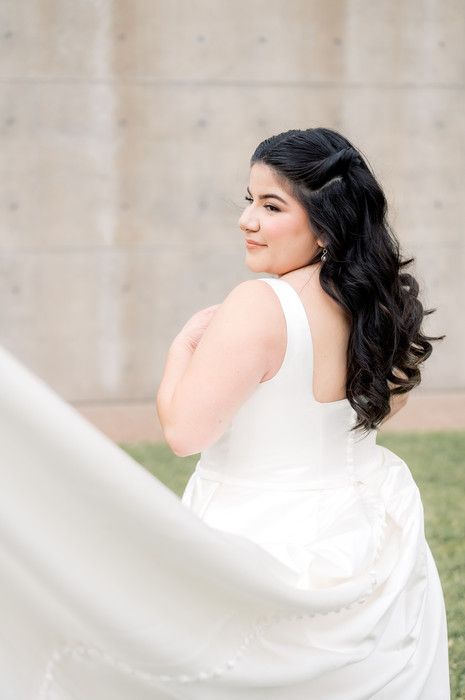 Bride in white gown with veil flowing, dark curly hair, smiling over shoulder outdoors.