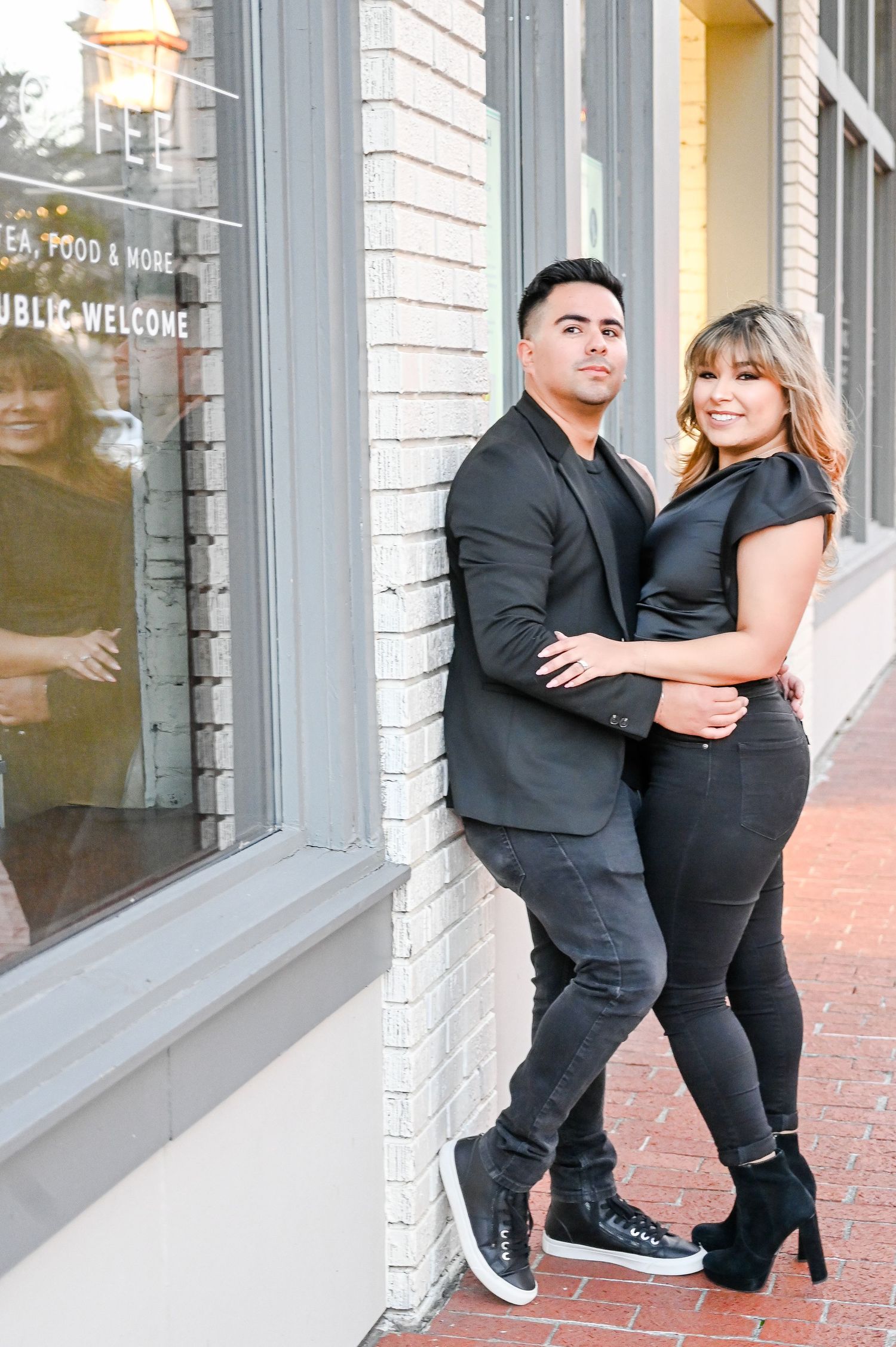 Stylish couple in all-black attire posing together against a brick building exterior on an urban sidewalk.
