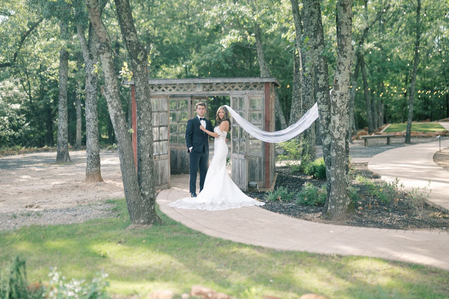 Bride and groom pose together under rustic wooden arbor surrounded by tall trees at outdoor venue.
