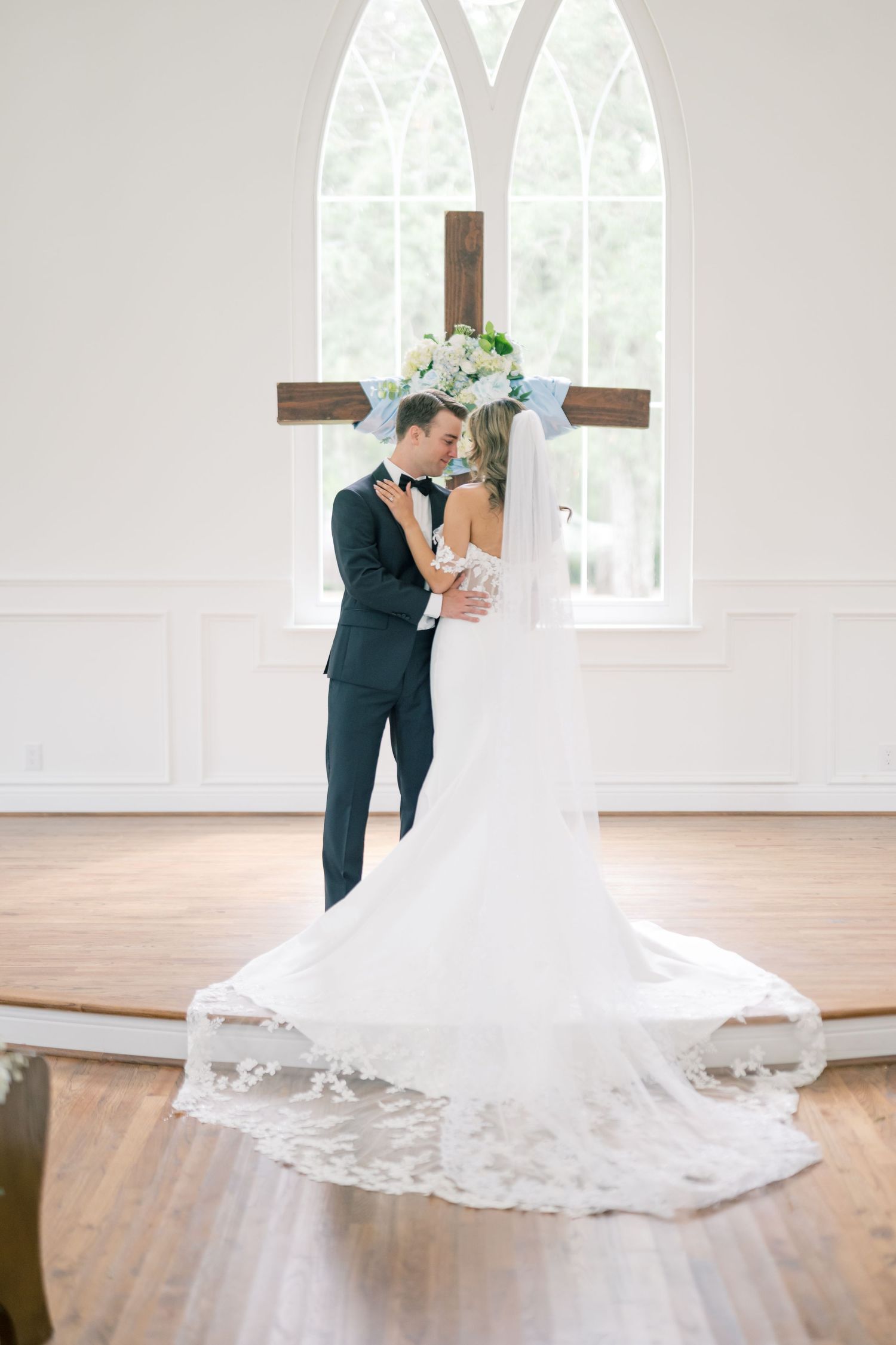 Bride and groom sharing a romantic embrace in a white chapel with arched windows and wooden cross.
