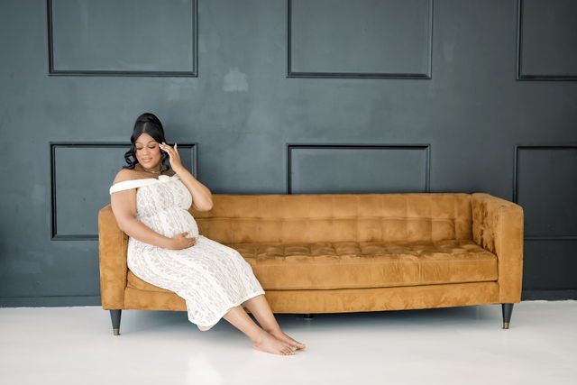 Pregnant woman in white lace dress sitting on a camel velvet sofa against a dark gray paneled wall.