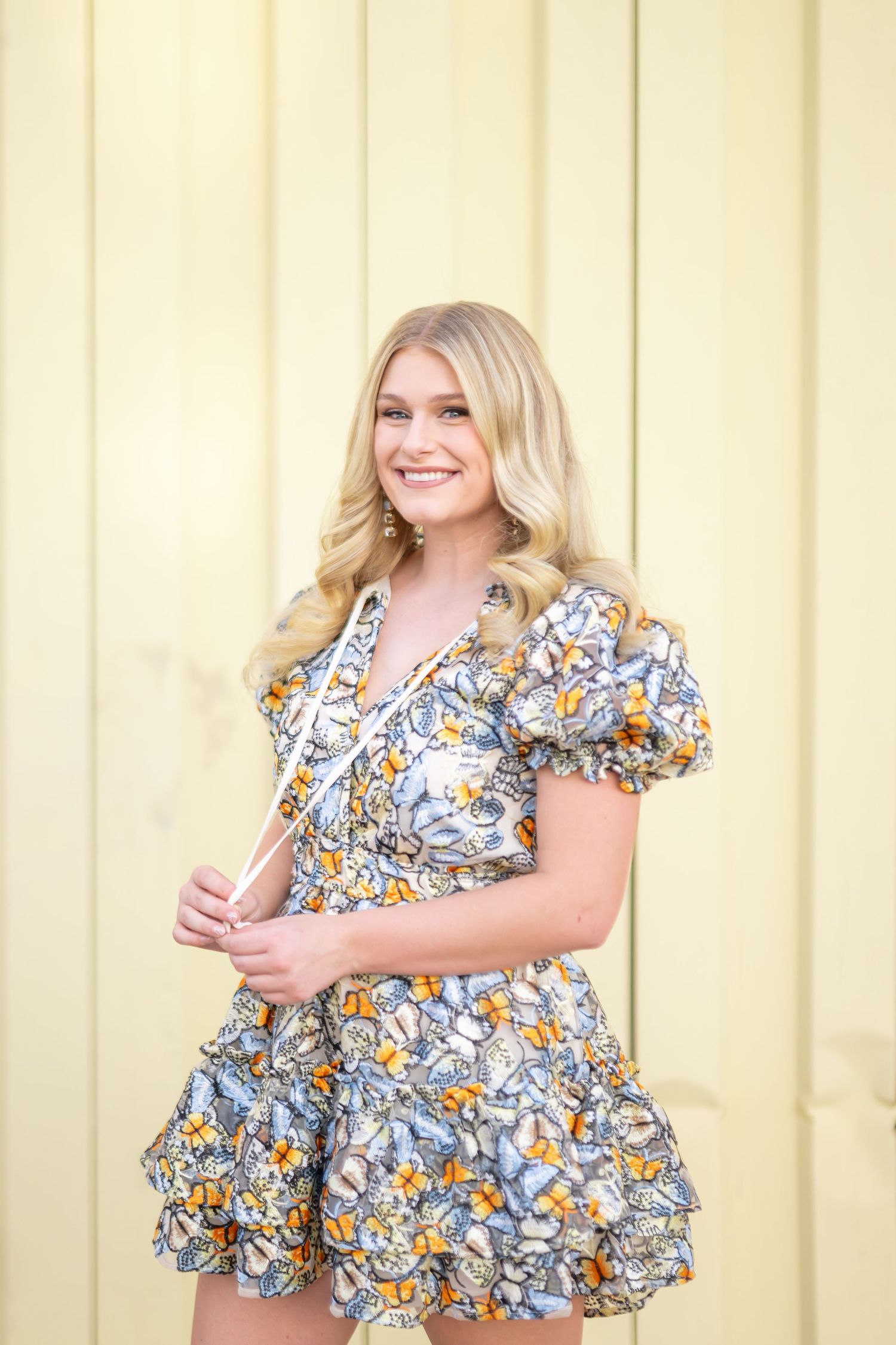 Smiling blonde woman in a floral blue and orange ruffled mini dress posing outdoors against a yellow wall.