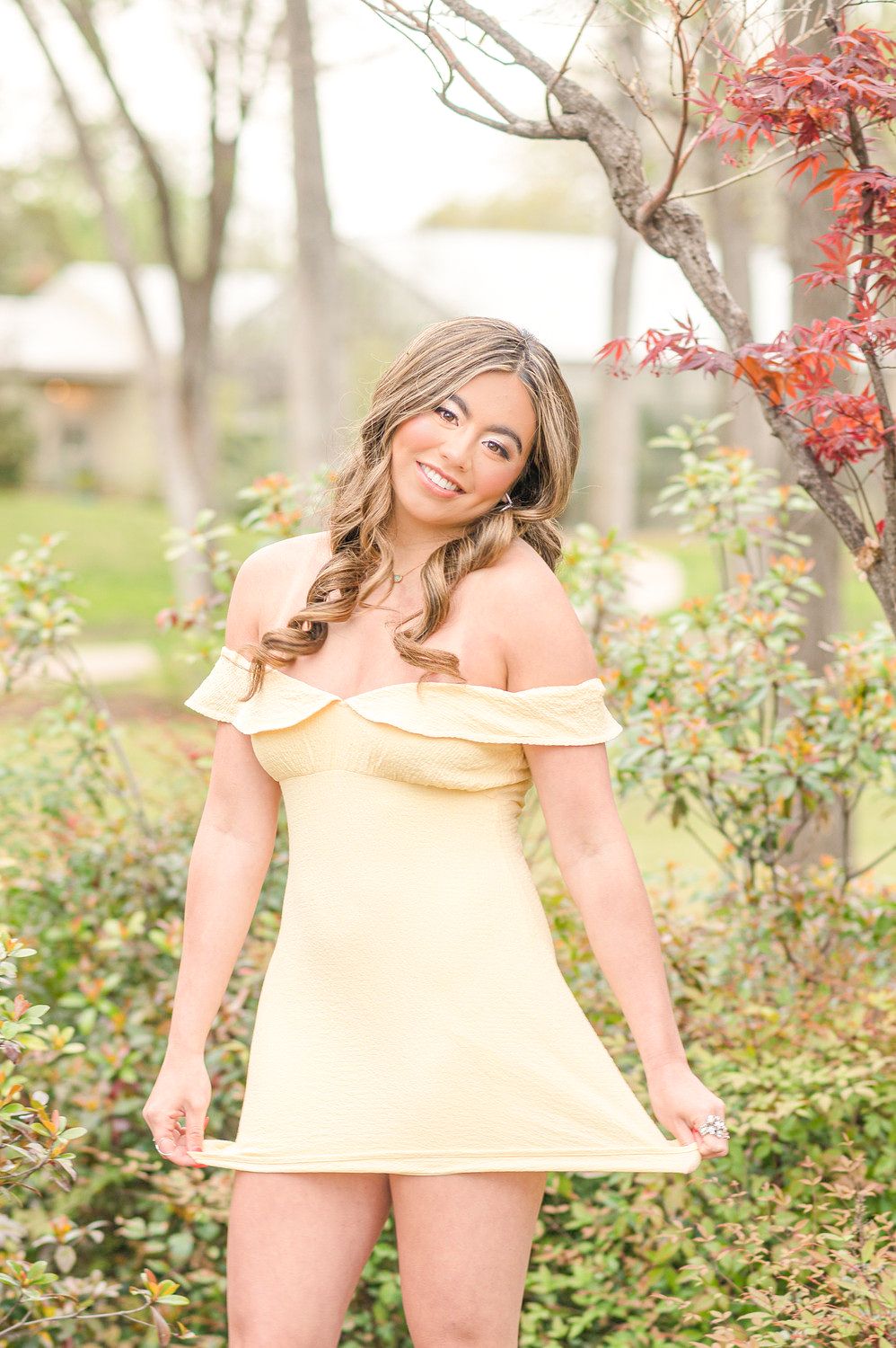 Smiling young woman in yellow off-shoulder mini dress poses among garden shrubs.