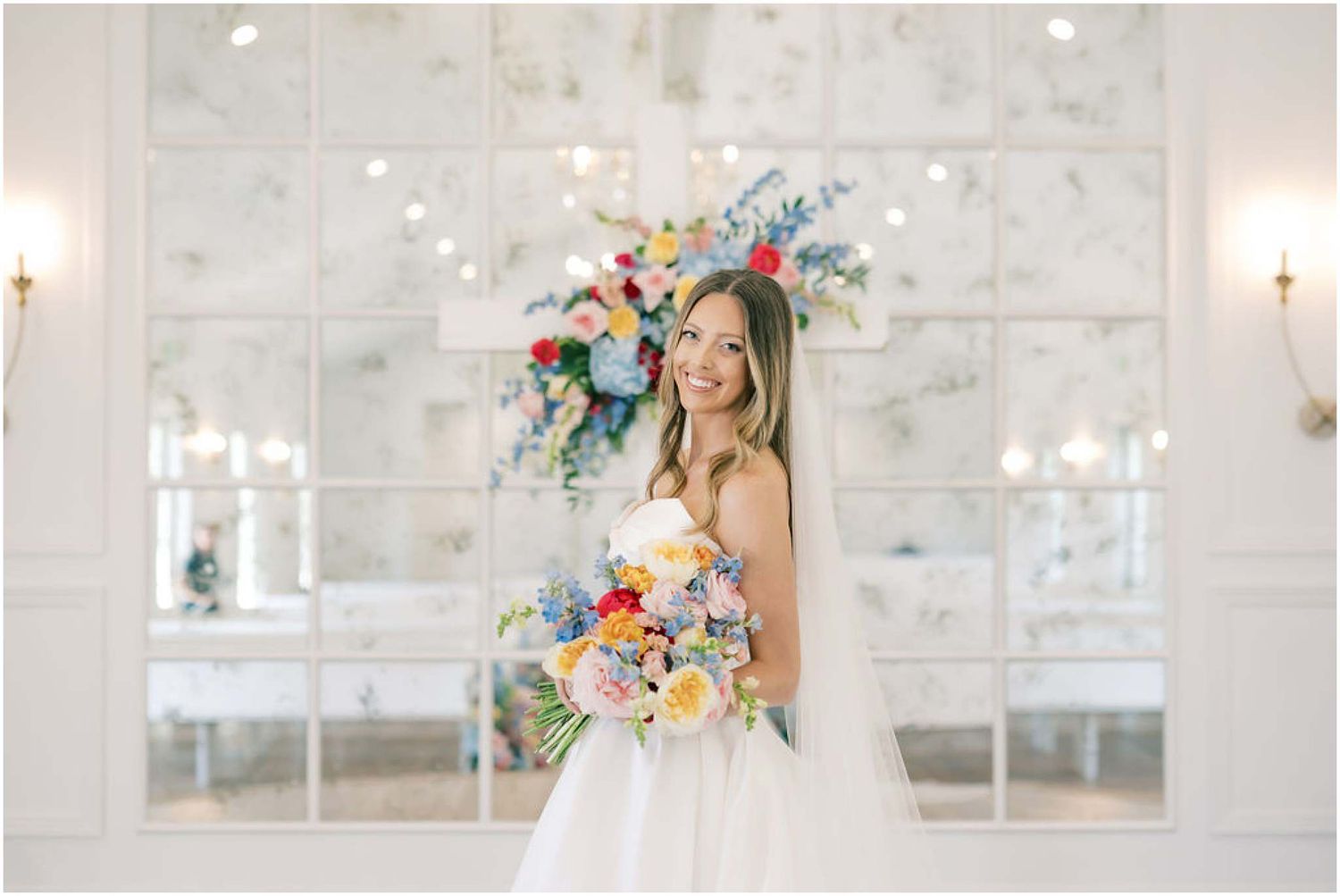 Bride in white gown holds colorful bouquet before mirrored wall with floral backdrop at elegant wedding venue.
