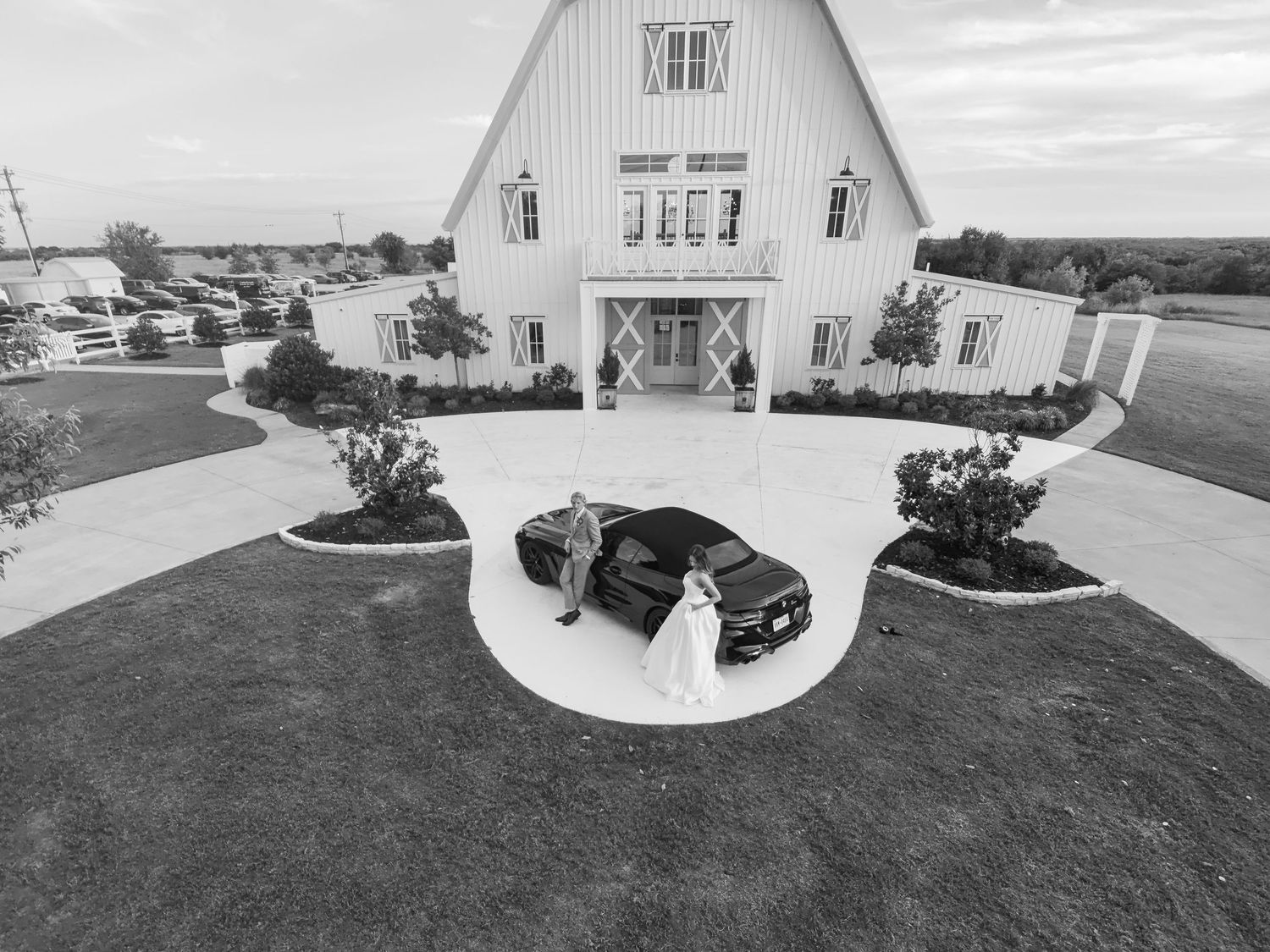 Black and white aerial photo of a bride and groom beside a dark car in front of a white barn wedding venue.