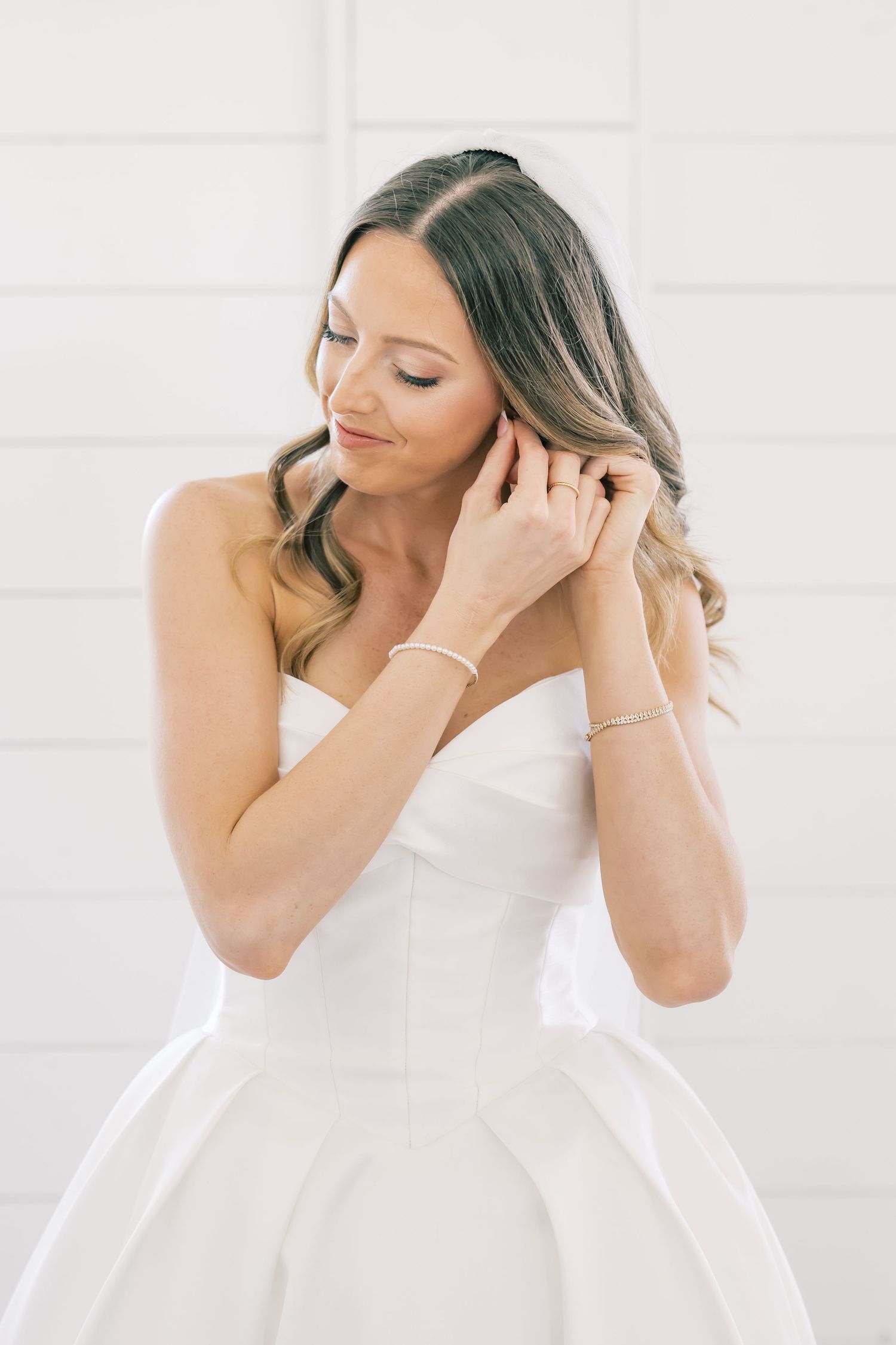 Bride in strapless white ball gown adjusting earring, with soft wavy hair and delicate bracelet.