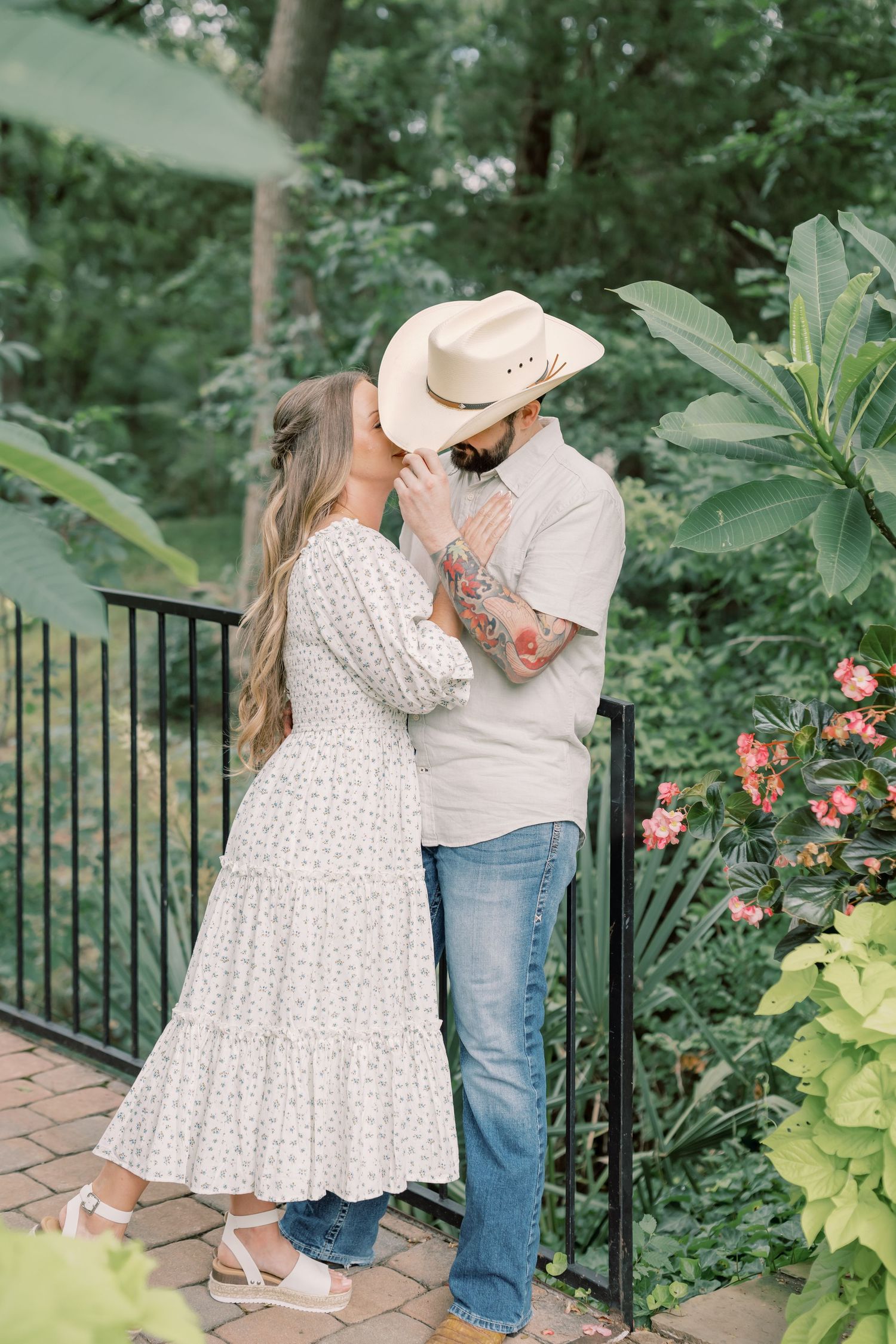 Couple sharing a kiss in a lush garden, man wearing a cowboy hat and tattooed arms visible.