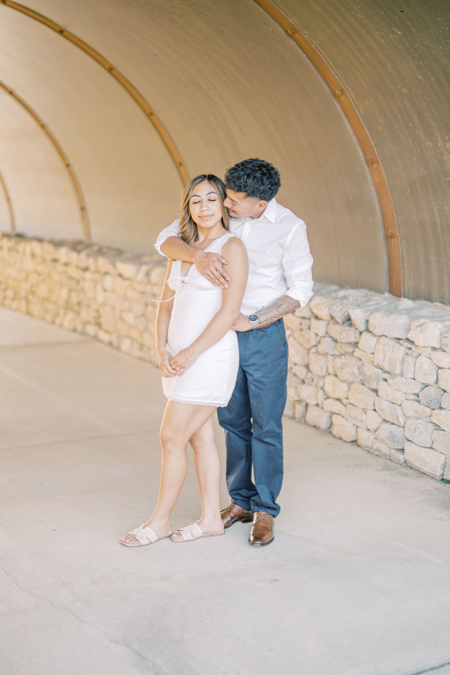 Couple embracing in a romantic tunnel setting, woman in white dress, man in white shirt and navy pants.