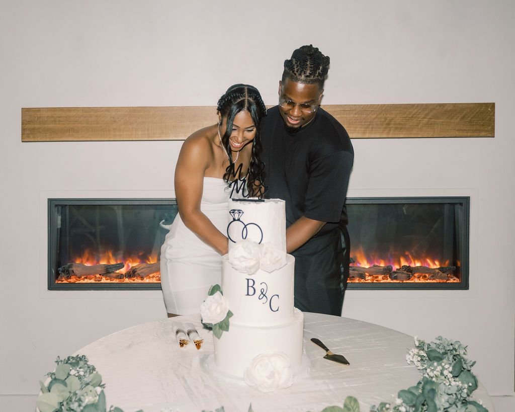 Couple cutting a white wedding cake with initials B&C near a modern fireplace with greenery decor.