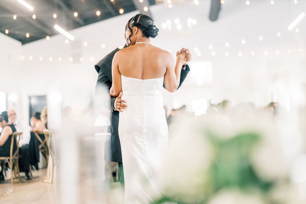 Bride and groom share their first dance at elegant wedding reception with string lights and floral arrangements.