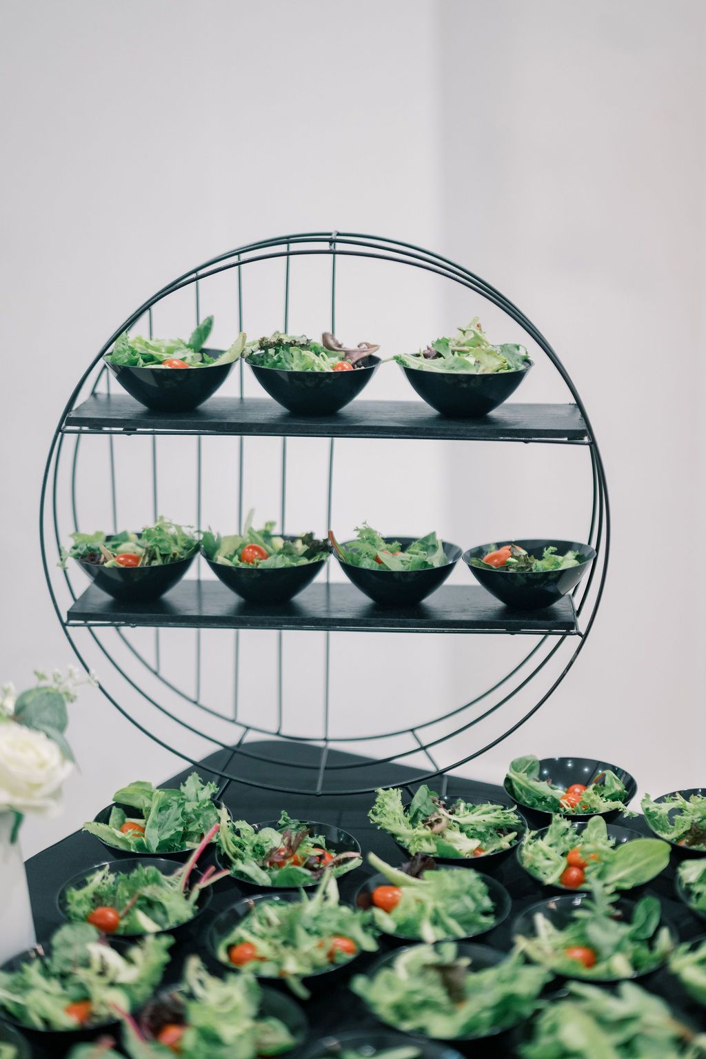 Circular metal display stand with three tiers holding black bowls of fresh green salads with cherry tomatoes at a catered event.