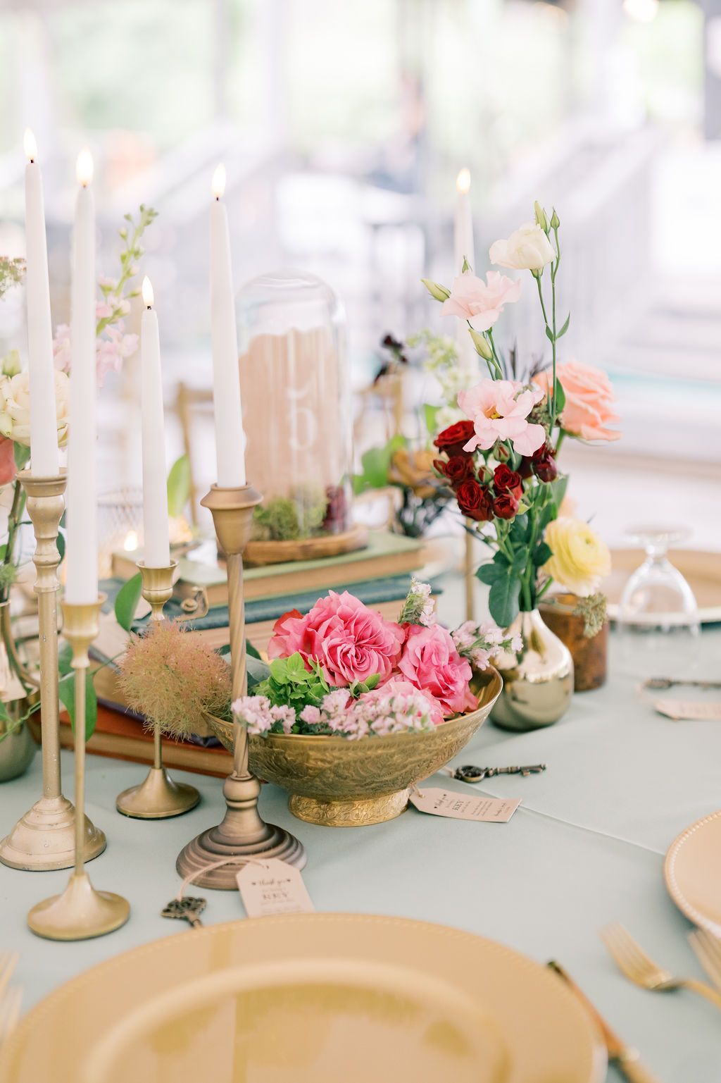 Elegant wedding table with pink roses in brass bowl, taper candles, and mixed floral arrangements on sage green linen.