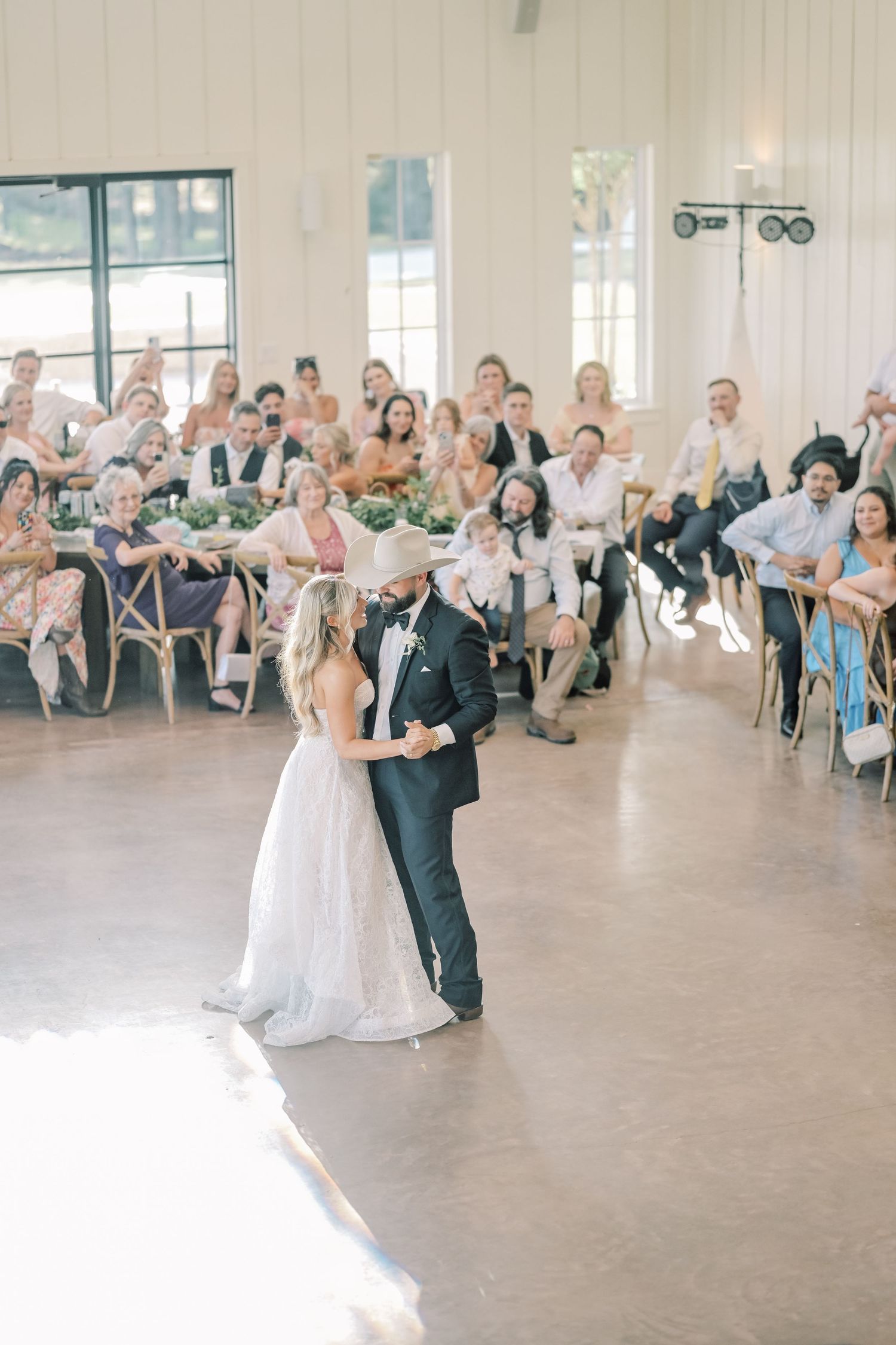 Bride and groom share first dance in bright reception hall surrounded by seated guests.
