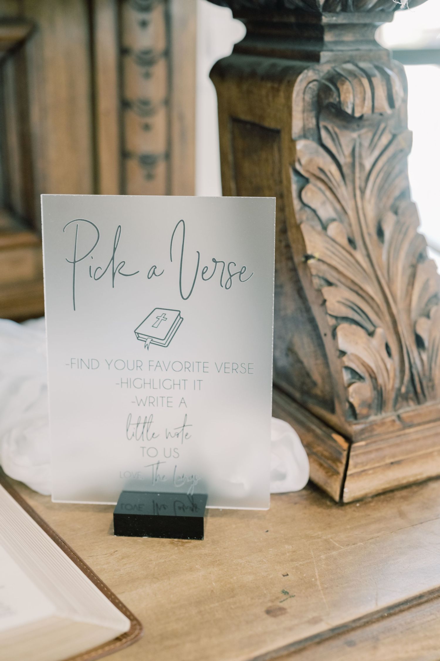 Elegant acrylic wedding sign reading Pick a Verse displayed on wooden table at reception.
