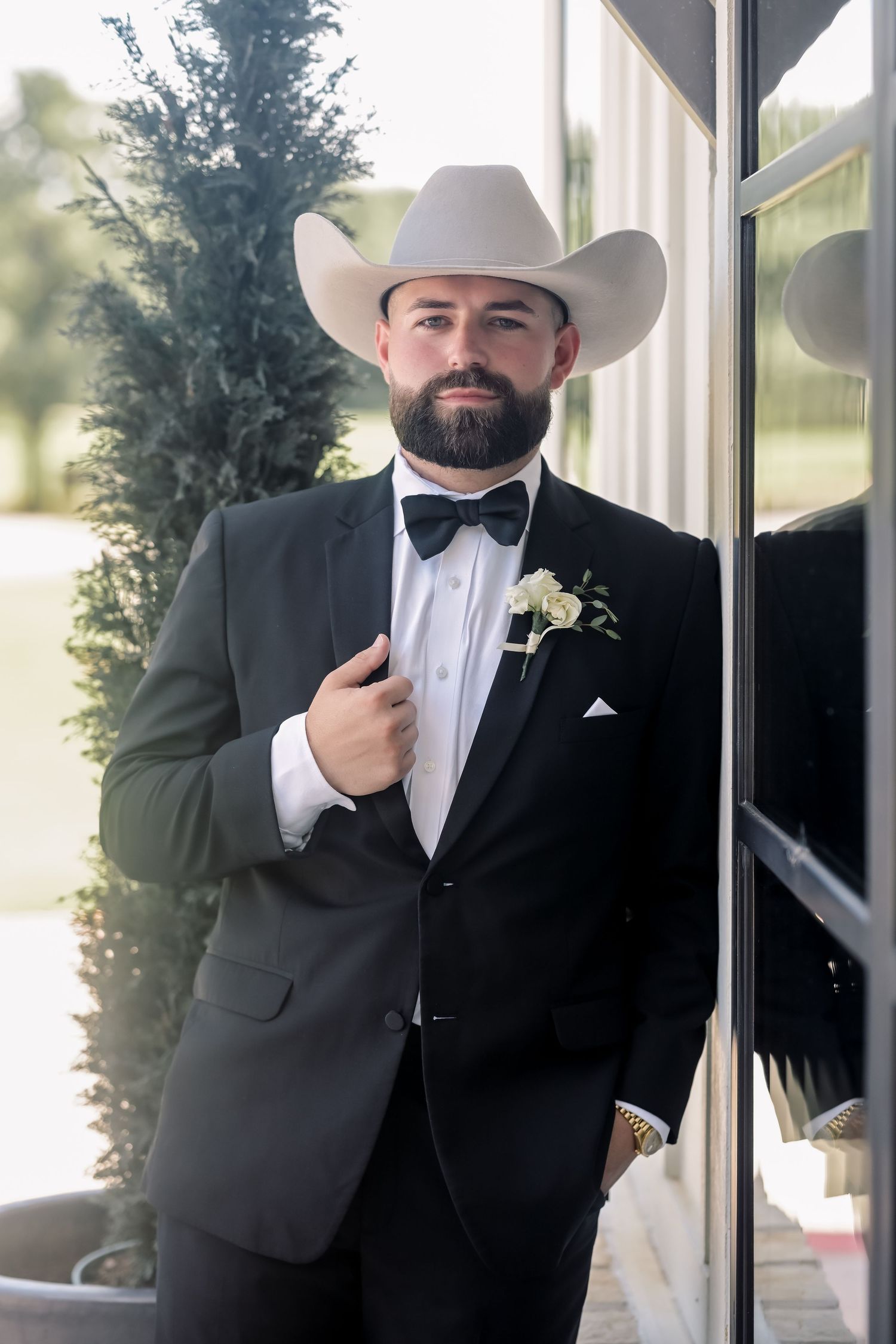 Dapper groom in black tuxedo and white cowboy hat poses with boutonniere before wedding.