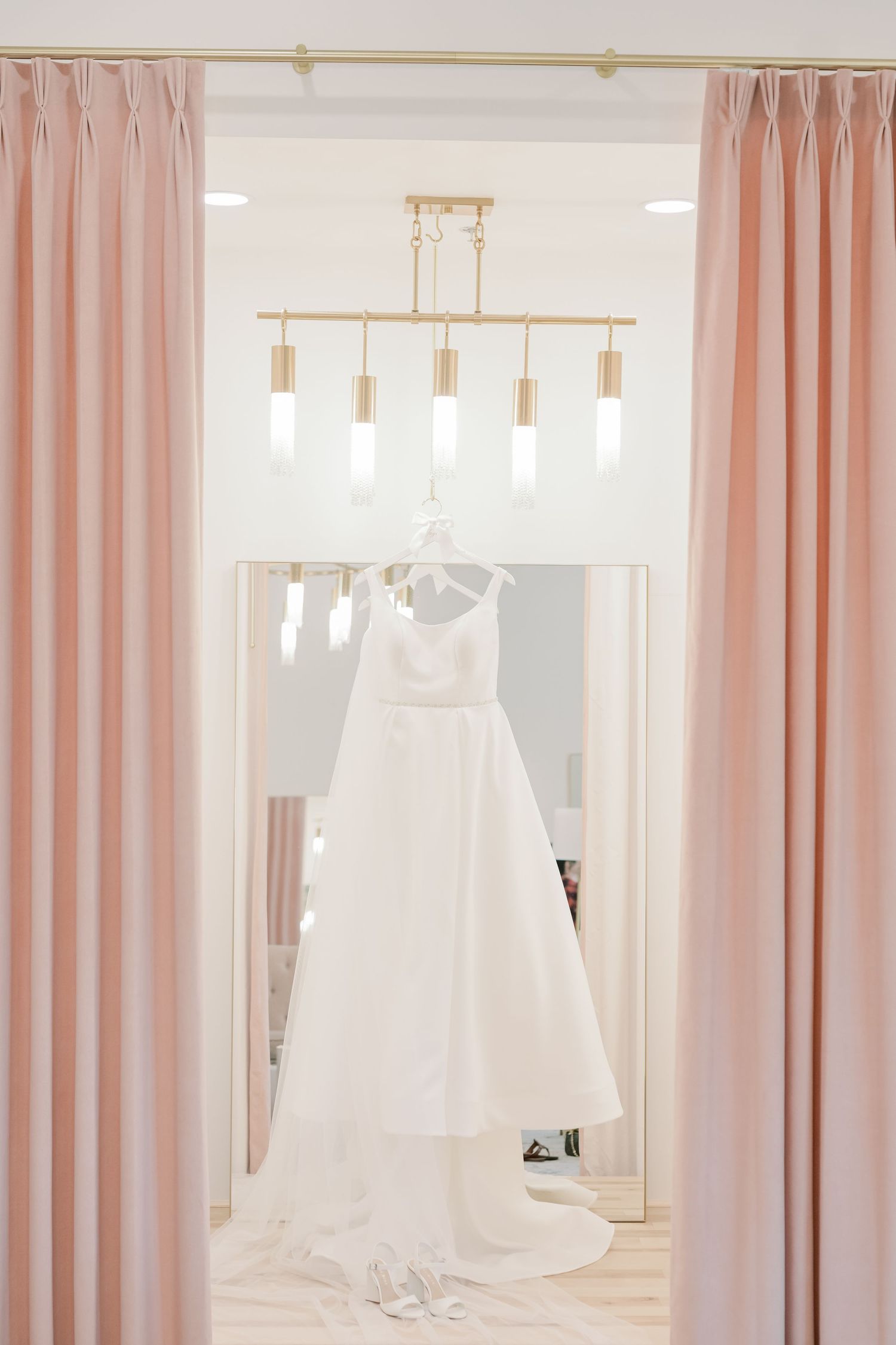 White wedding gown hanging from a gold chandelier framed by soft pink curtains in a bridal boutique.
