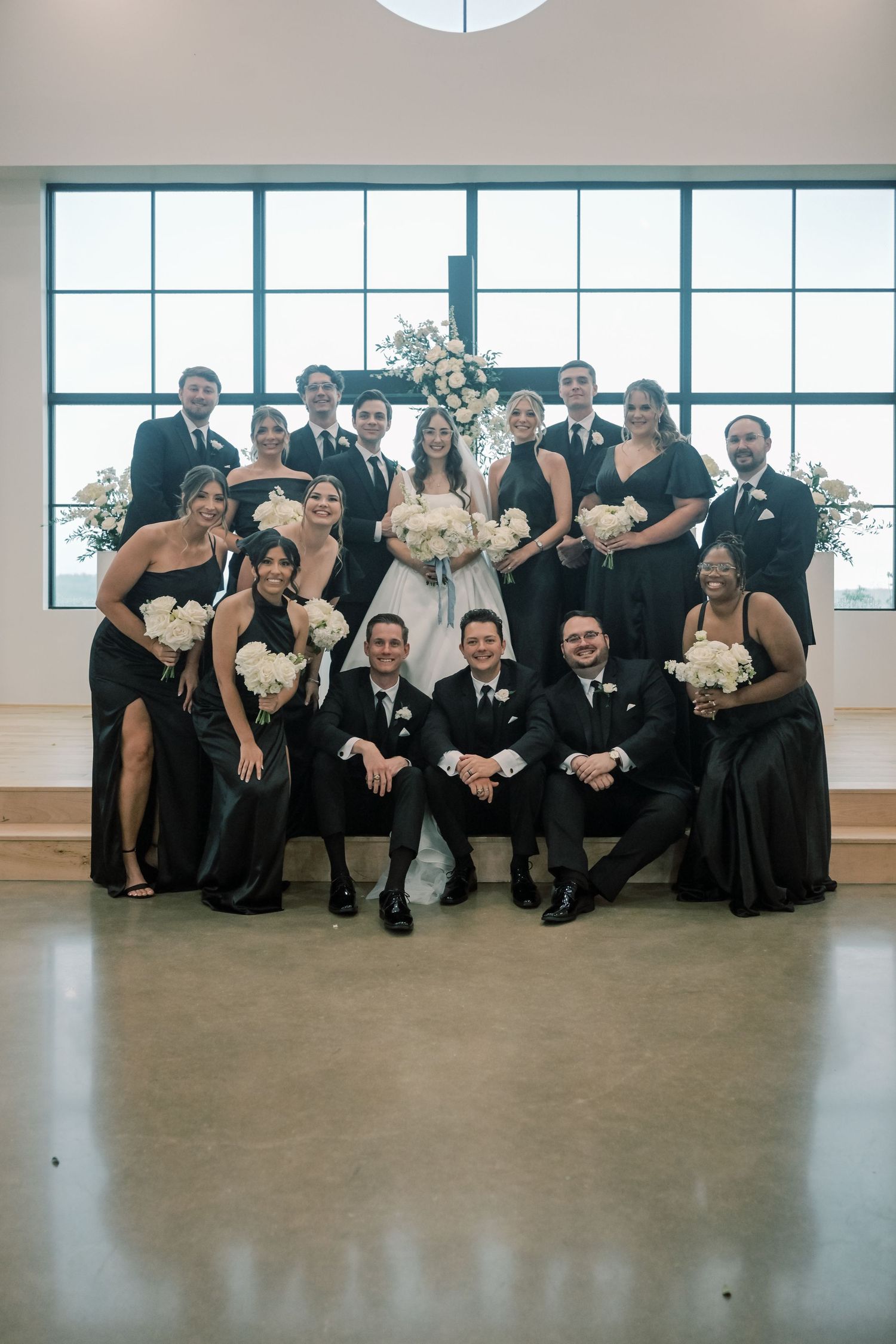 Wedding party in black formal attire posing with bride and groom holding white floral bouquets in bright venue.