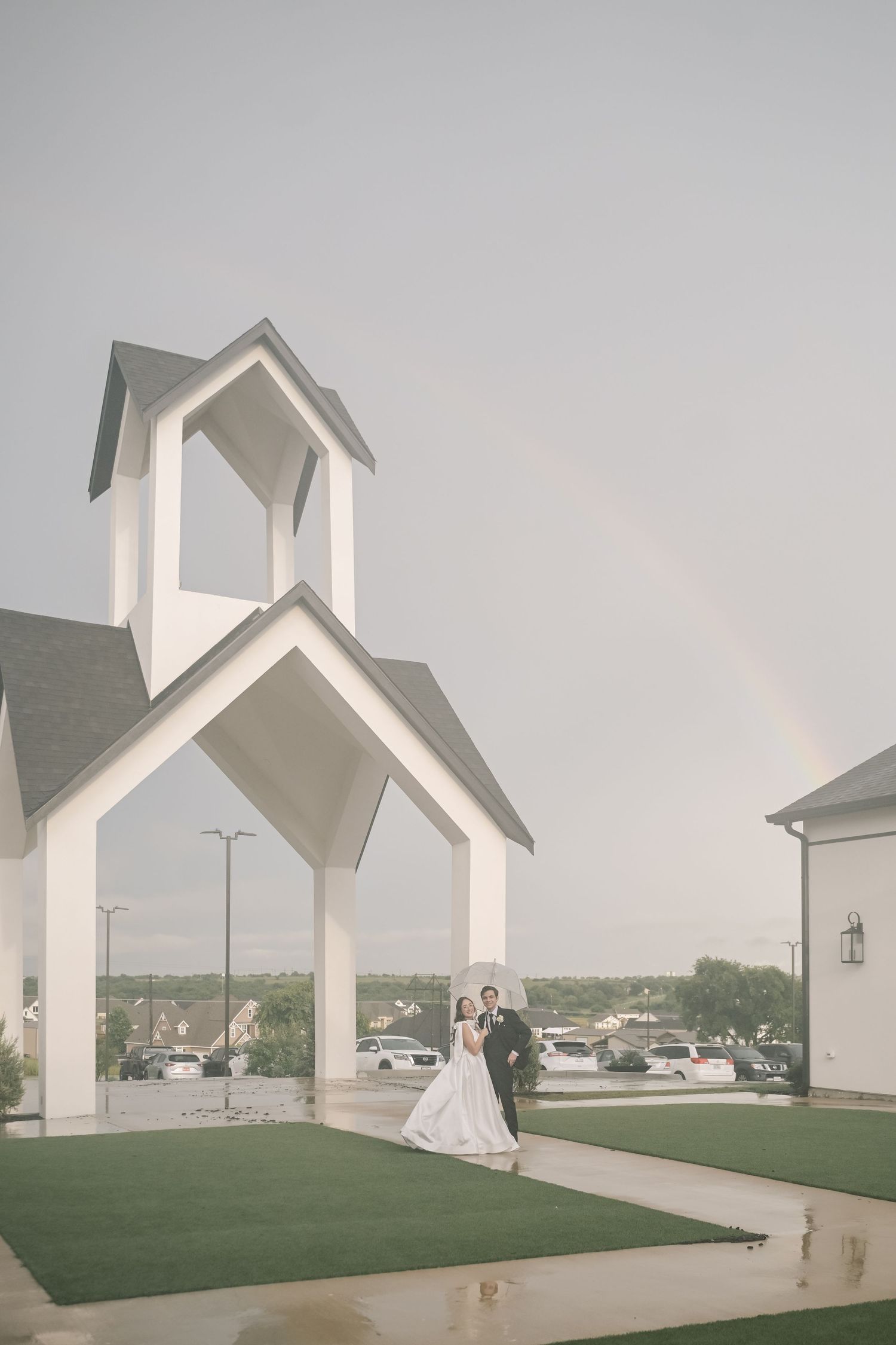 Bride and groom pose under white chapel arch with rainbow in background after rainy wedding.