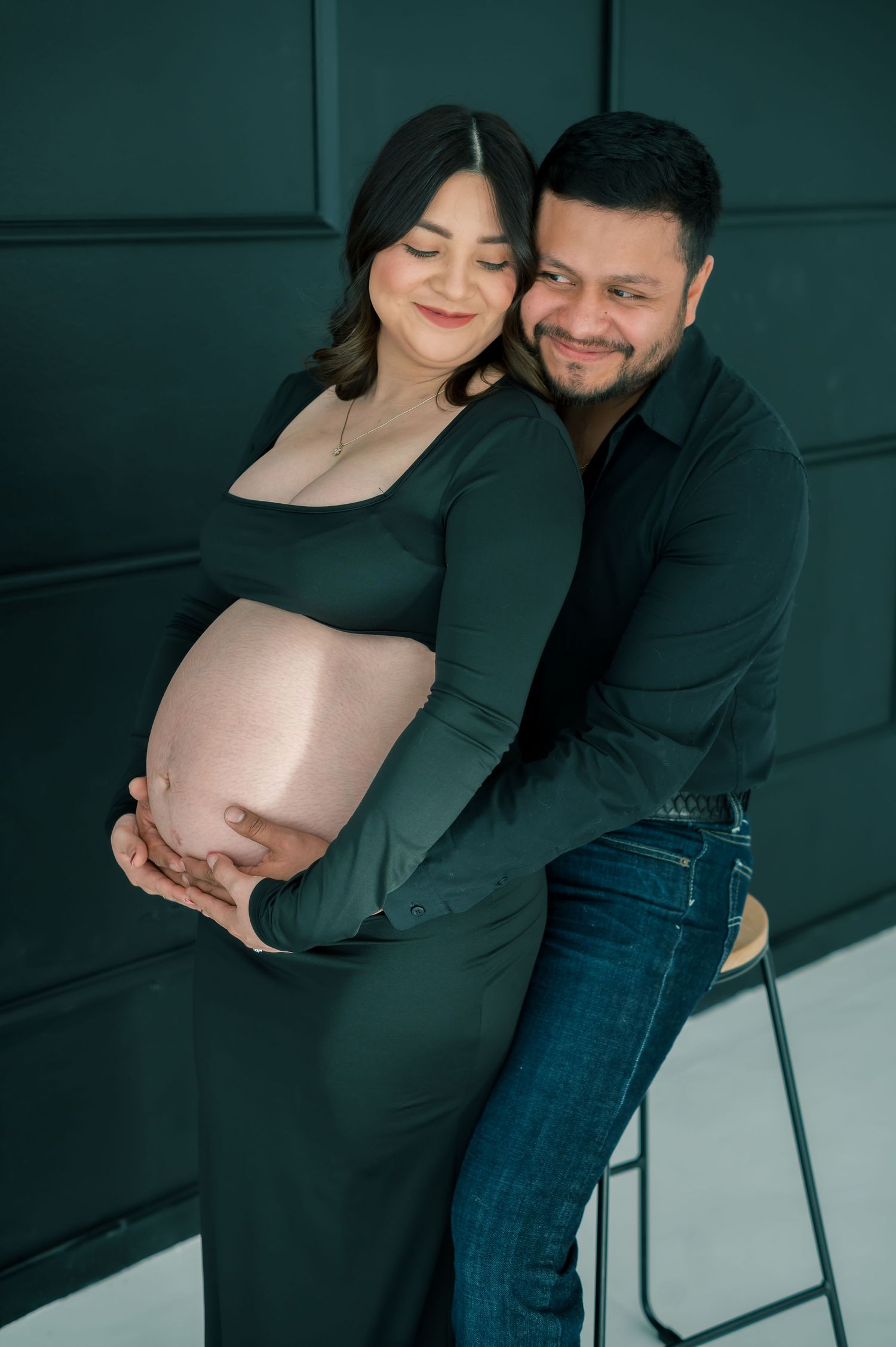 Couple sharing tender moment in maternity shoot, man smiling while embracing pregnant woman in dark green dress.