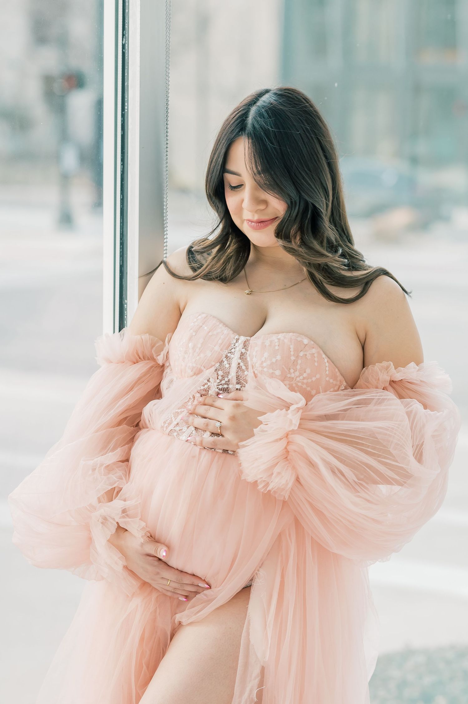 Smiling pregnant woman in blush tulle maternity robe gently holding her baby bump.