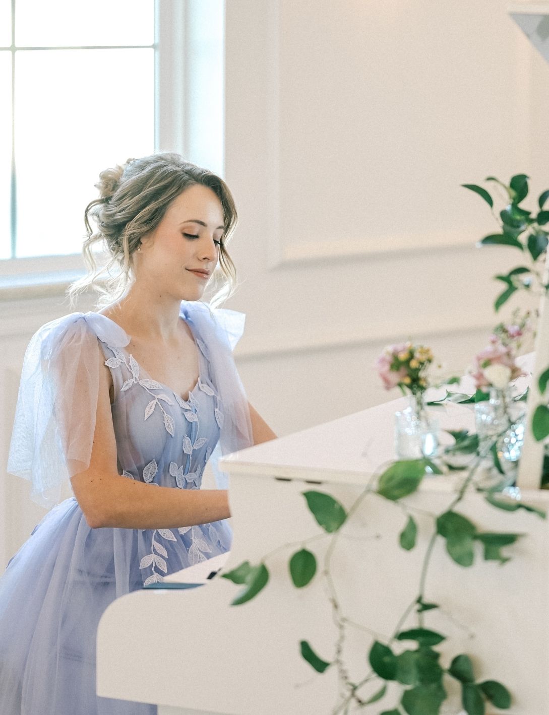 A bride in a light blue tulle gown with floral embroidery plays a white piano in a bright elegant room.