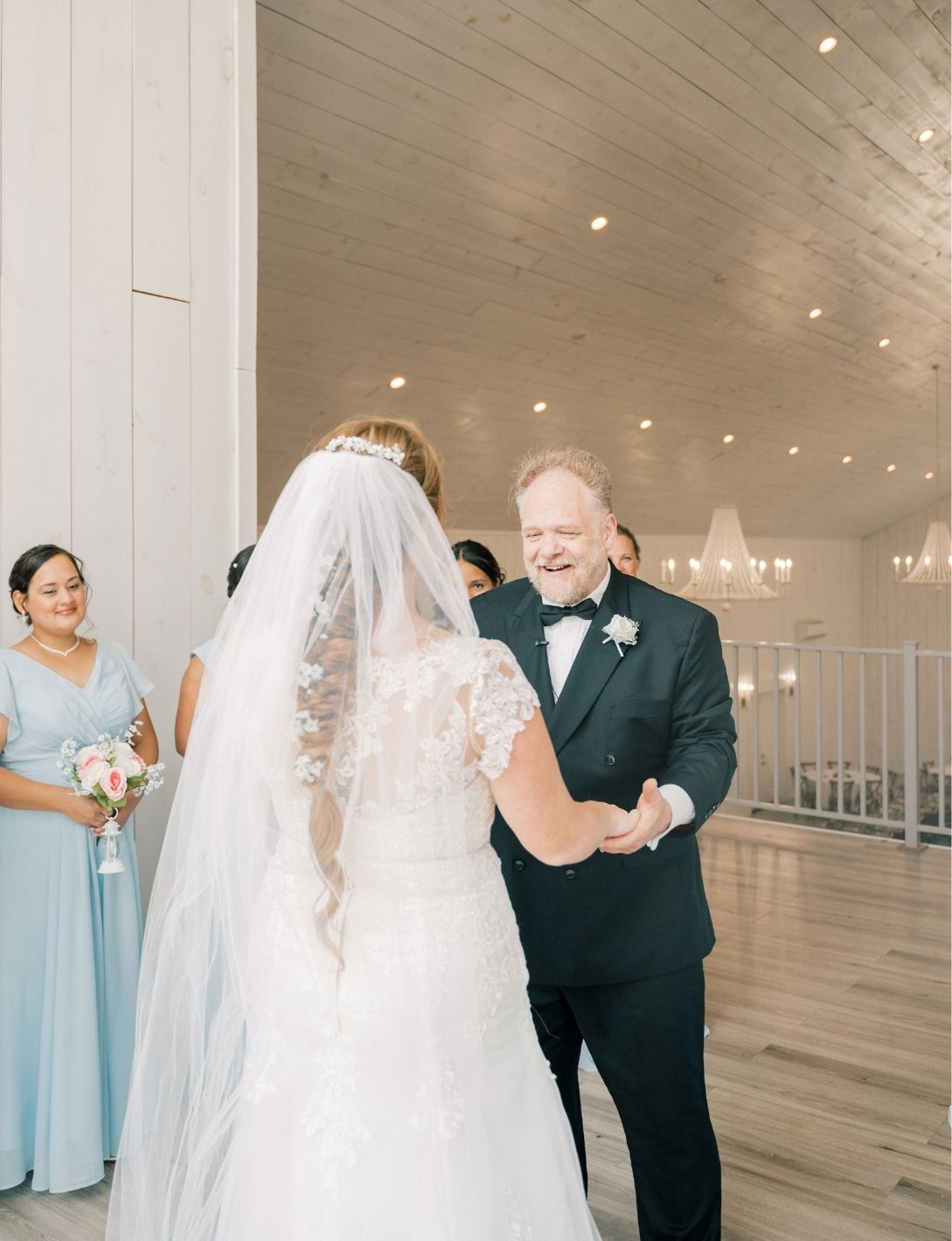 Father smiling at bride in white lace gown and veil during emotional first look at elegant wedding venue.