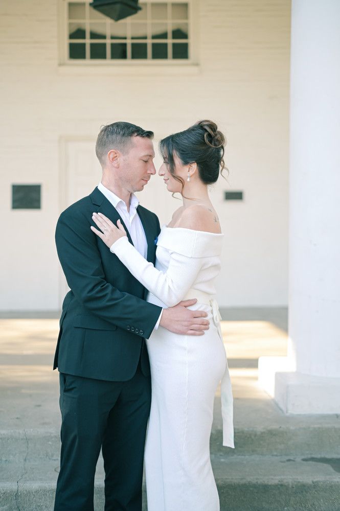 Bride and groom sharing romantic moment, she in elegant off-shoulder white dress, he in dark suit, nearly kissing.