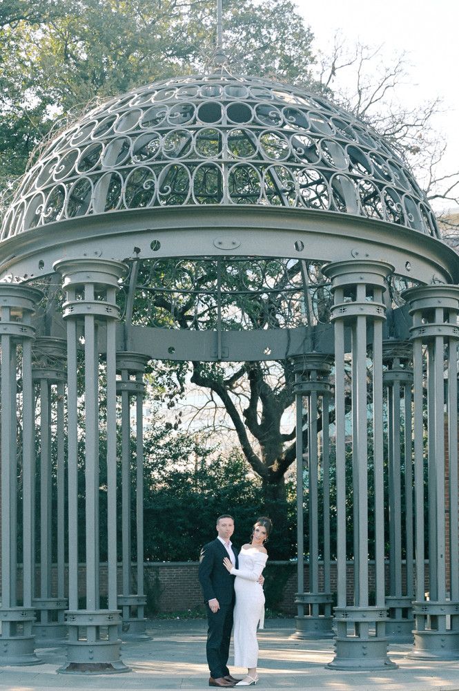 Couple poses romantically under an ornate iron gazebo with a domed lattice roof in a park setting.