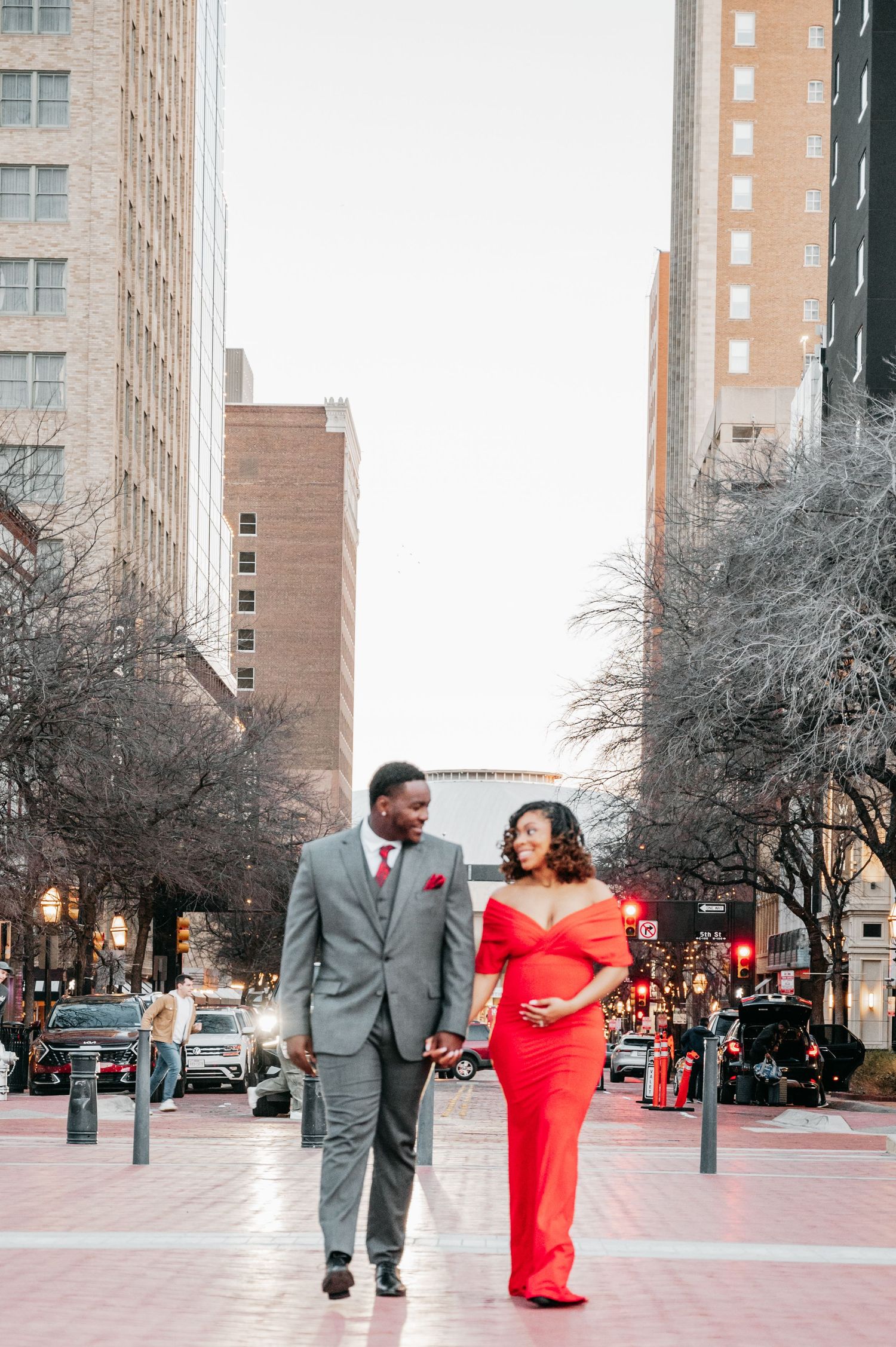 Couple walking hand-in-hand on city street, woman in red maternity gown, man in gray suit.