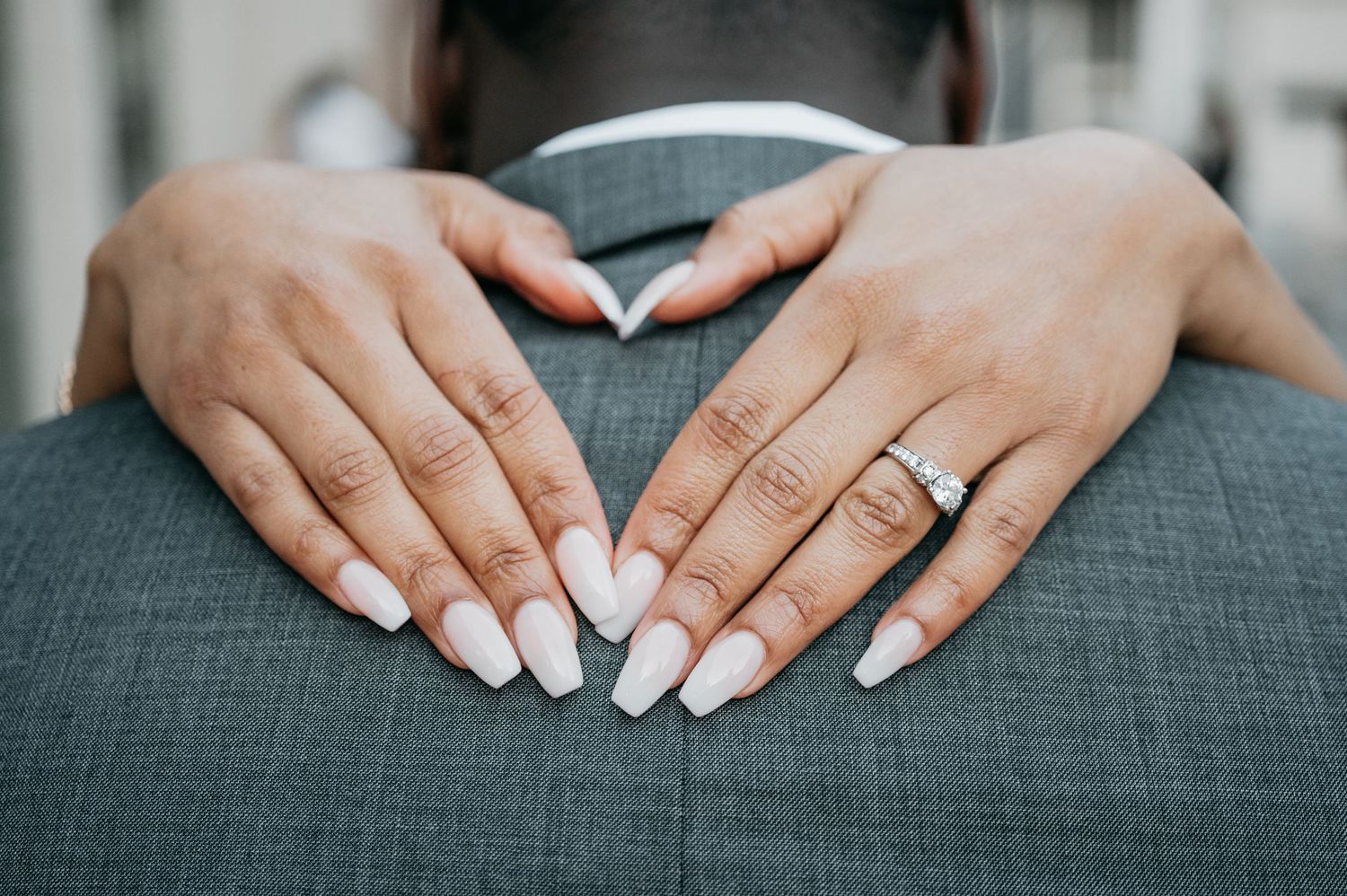 Close-up of hands with white almond nails and diamond engagement ring resting on a gray suit jacket.