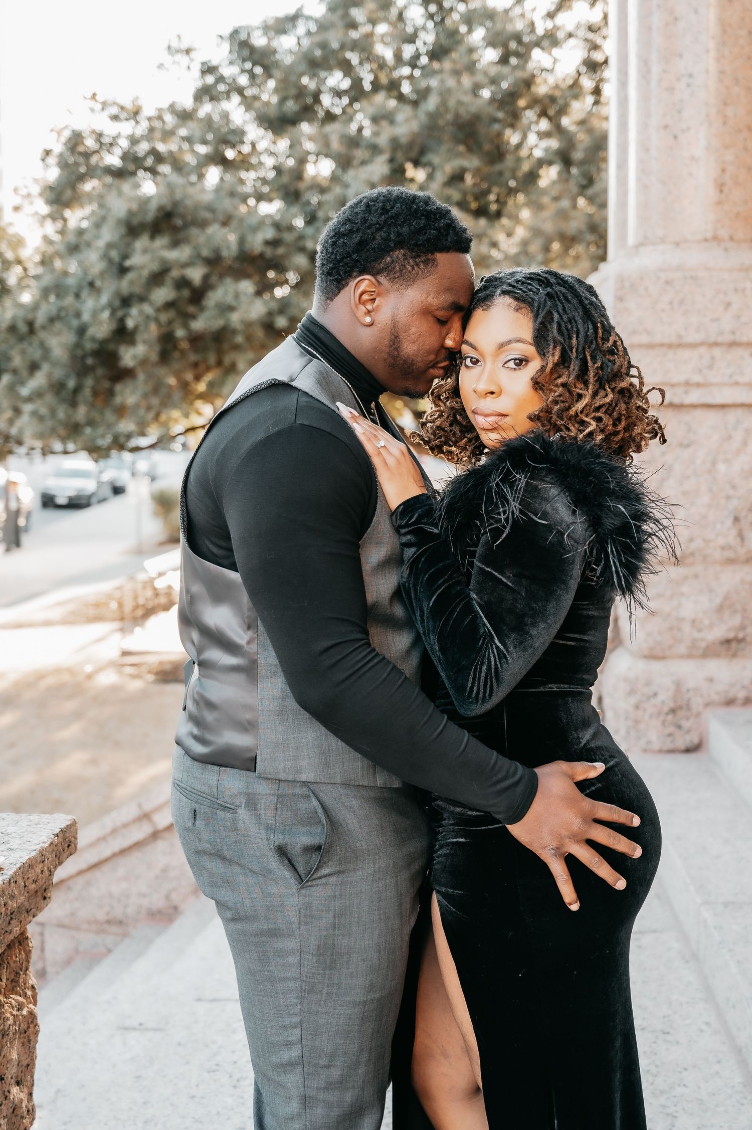 A couple in elegant black and gray formal attire share a romantic embrace outdoors near a stone column.