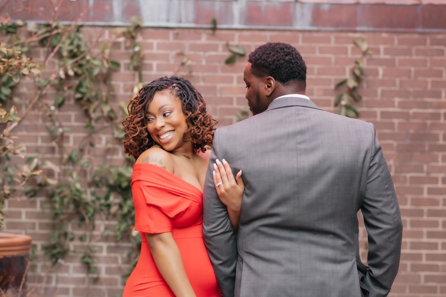 A smiling woman in a red off-shoulder dress stands with a man in a gray suit near a brick wall.
