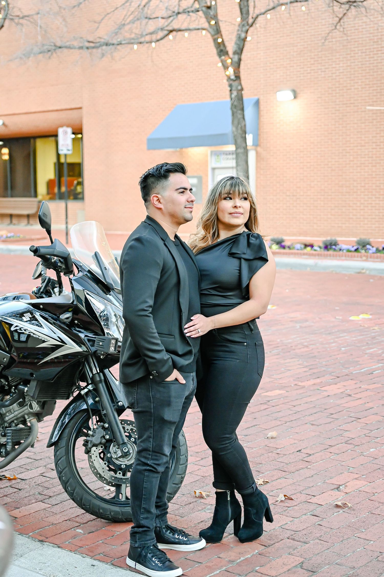 Stylish couple dressed in all black standing together near a parked motorcycle outdoors.