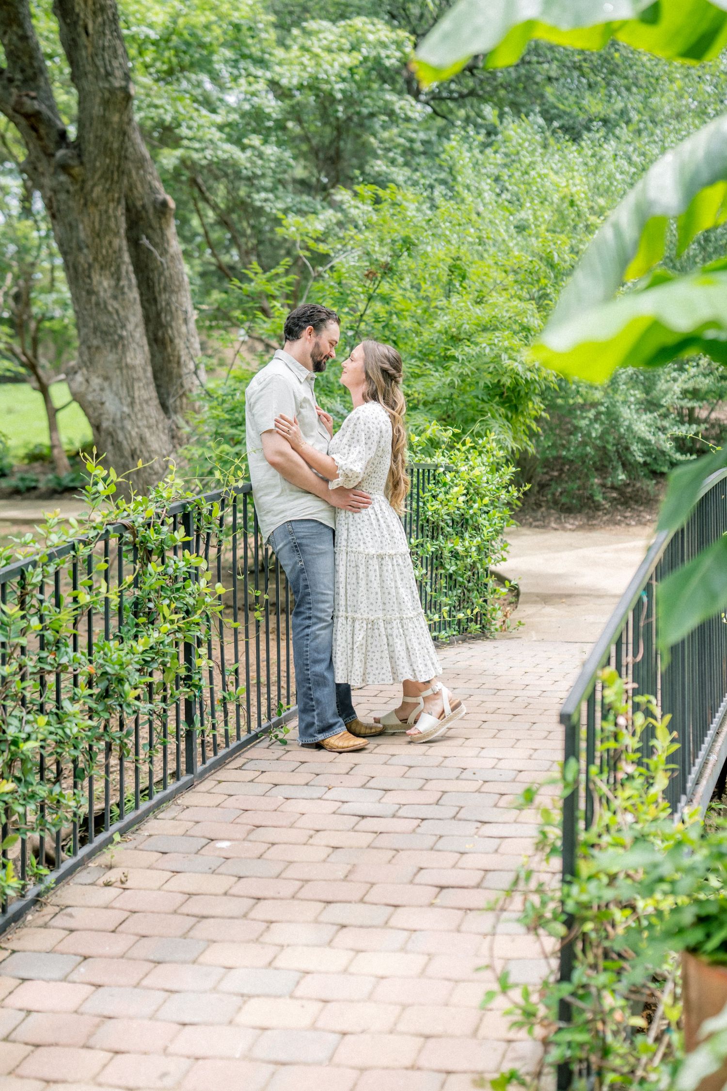 Couple embracing on a brick garden path surrounded by lush greenery, sharing a romantic moment near black iron railings.