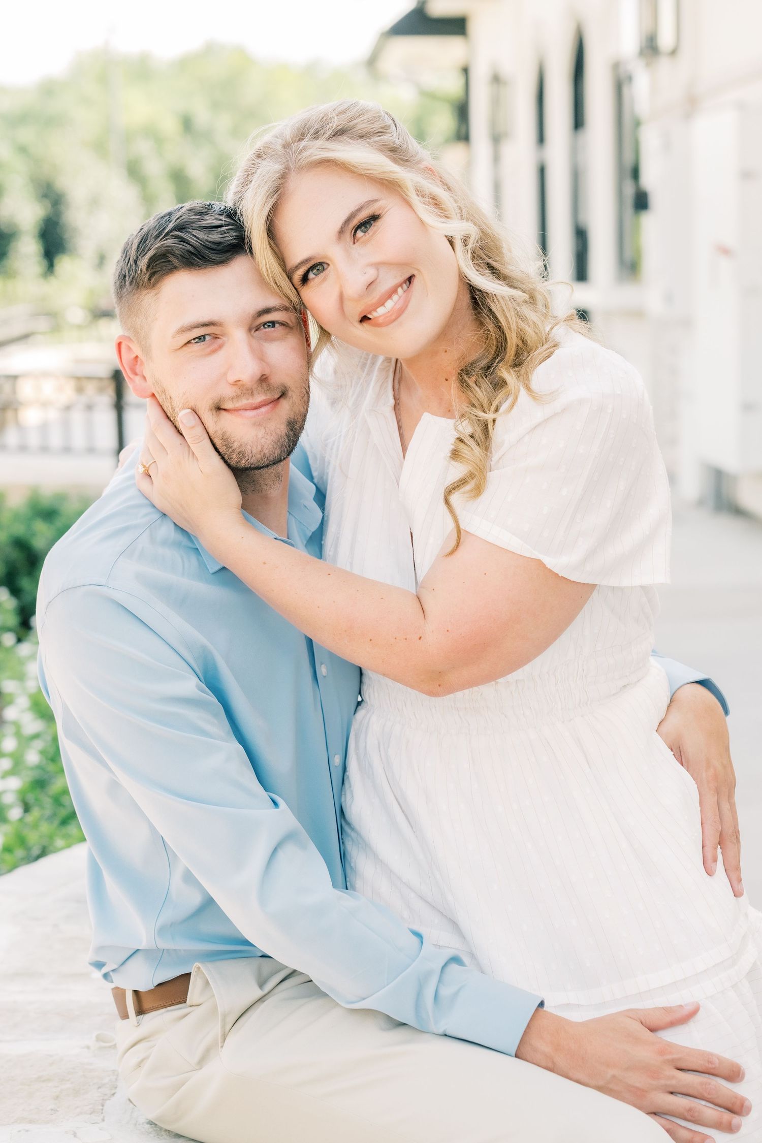 Smiling couple in light blue and white outfits embracing outdoors near a classic building facade.