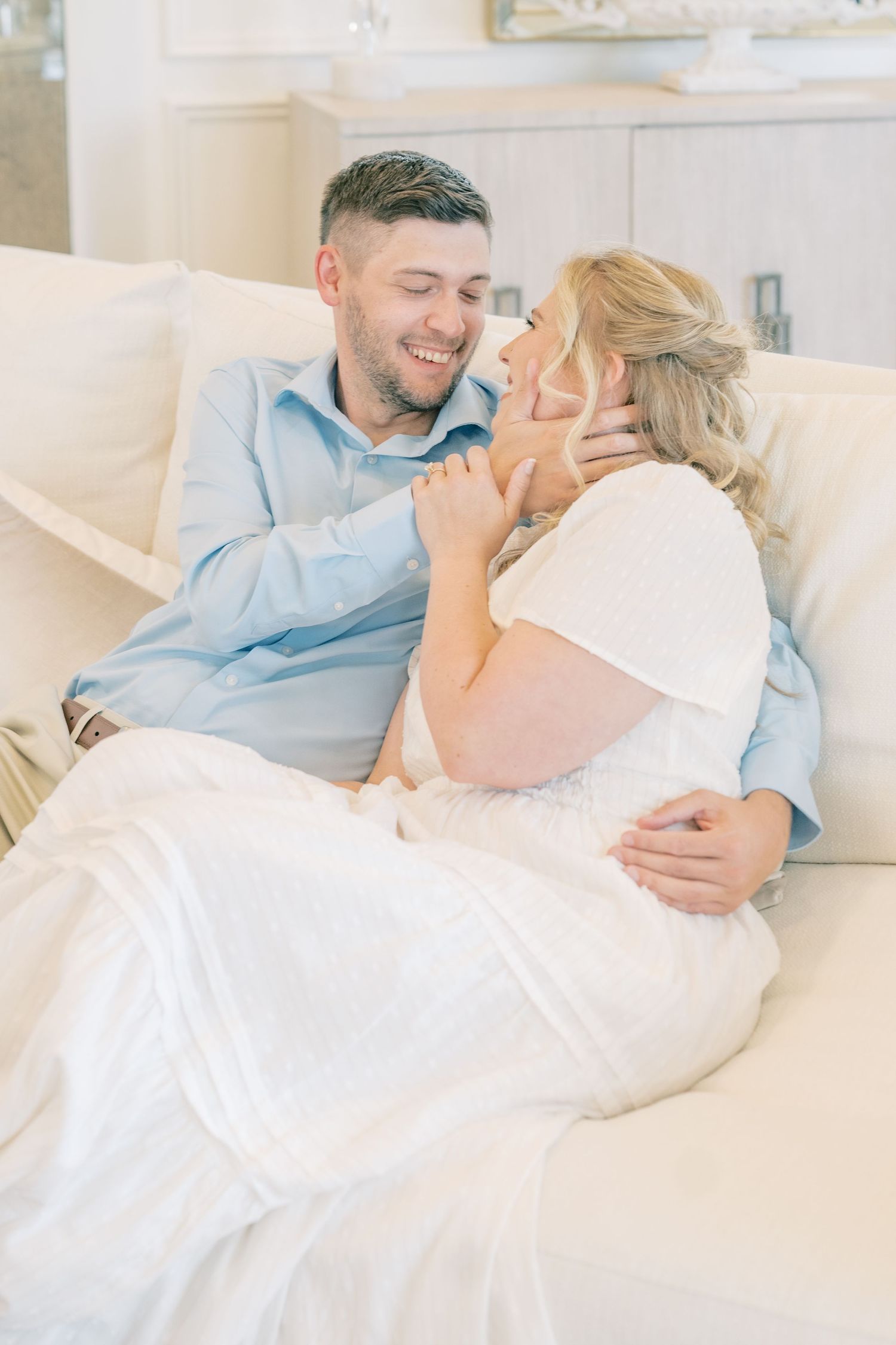 A smiling couple cuddles on a cream sofa, the man in a blue shirt gently touching the woman's face.