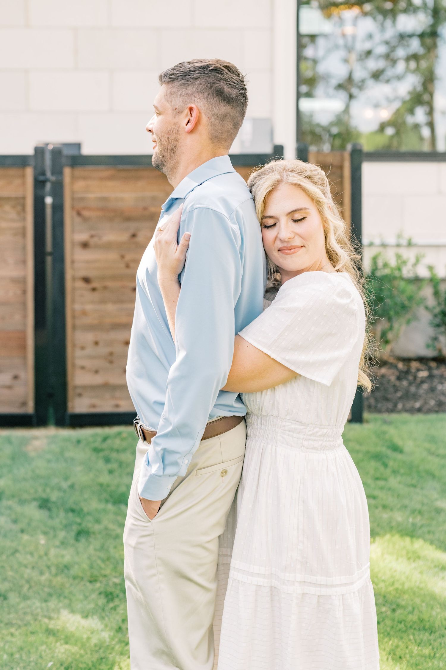 Woman in white dress hugging man in light blue shirt from behind while smiling outdoors near a wooden fence.