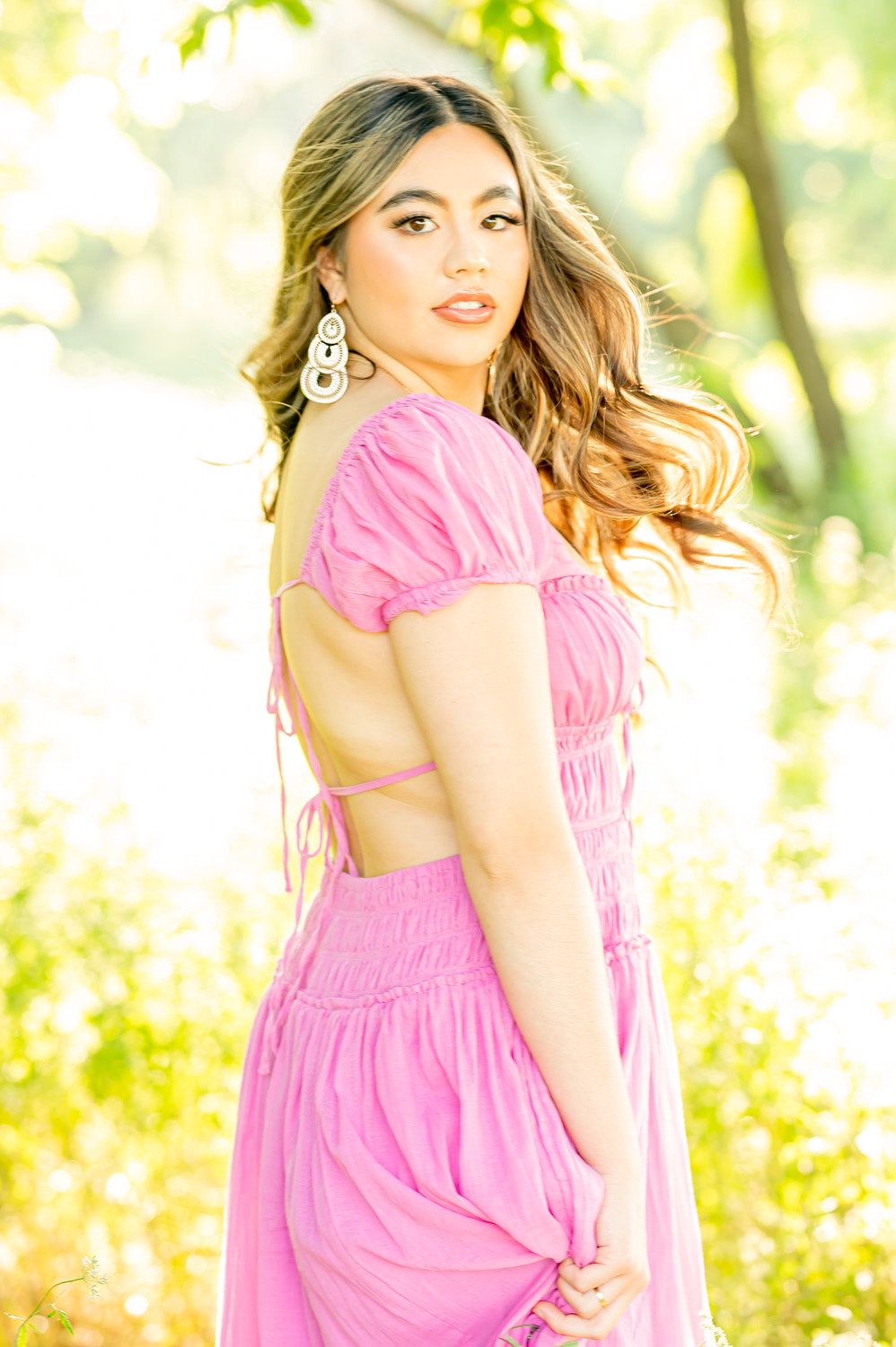 Young woman in a pink backless dress with statement earrings poses outdoors in golden sunlight.