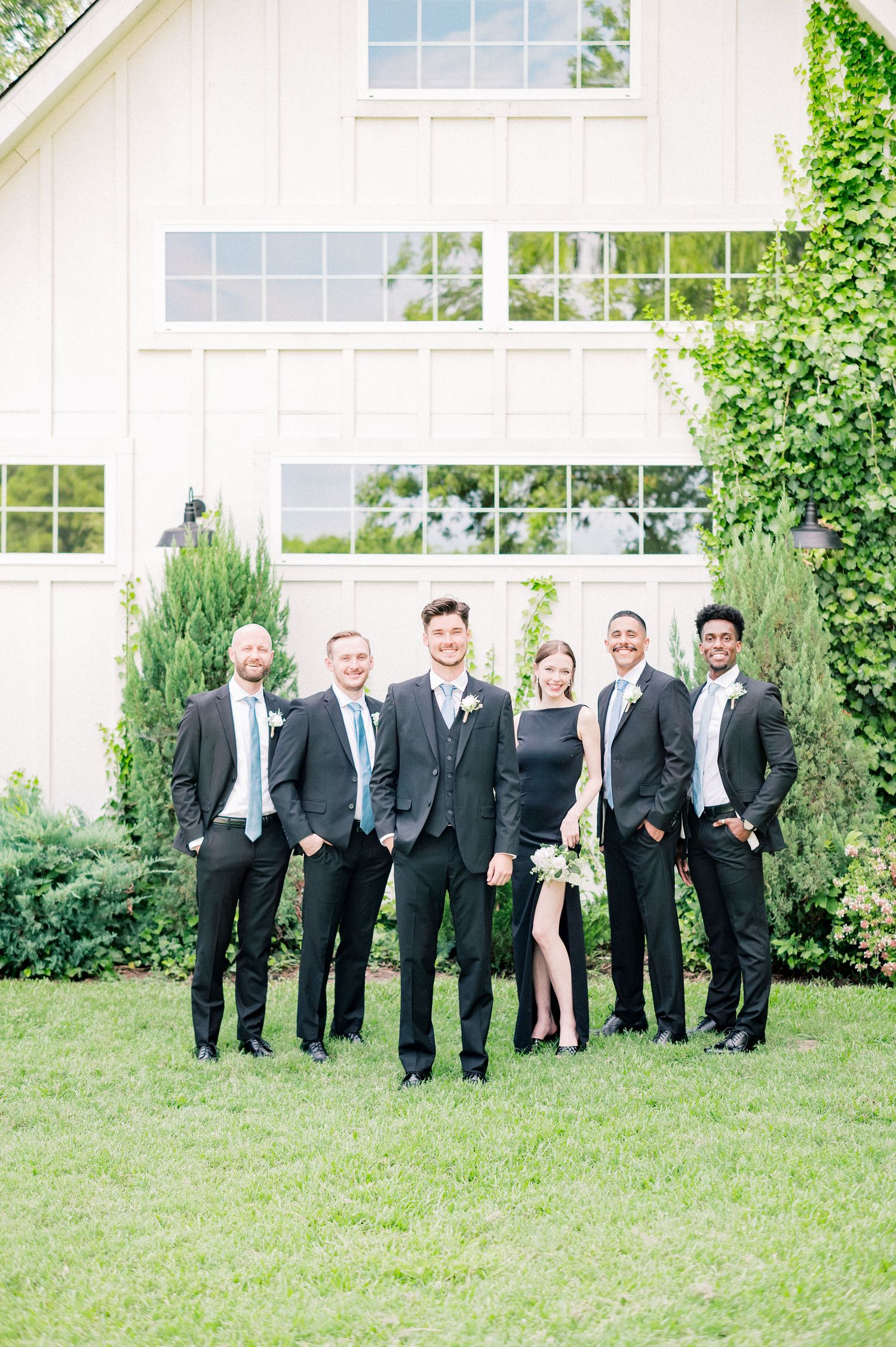 Groomsmen in black suits pose casually on grass outside a white building.