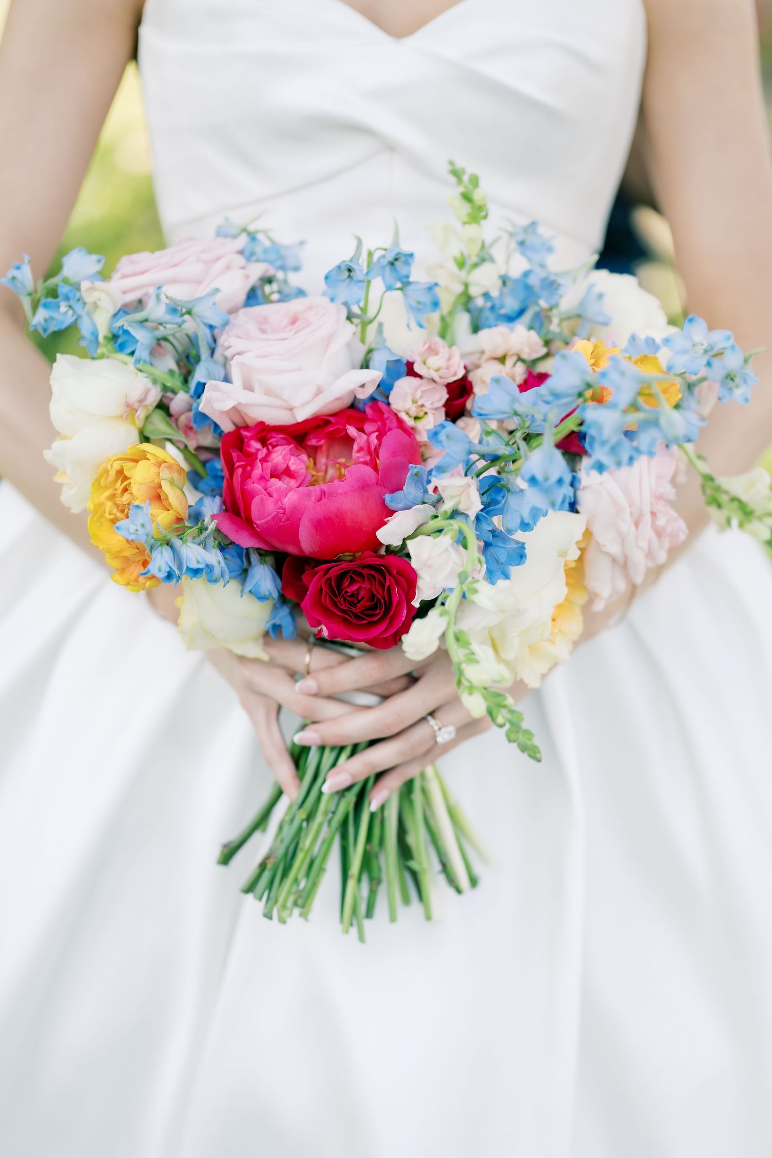 Bride holding a vibrant wedding bouquet with pink peonies, red roses, blue delphiniums, and yellow flowers in white dress.