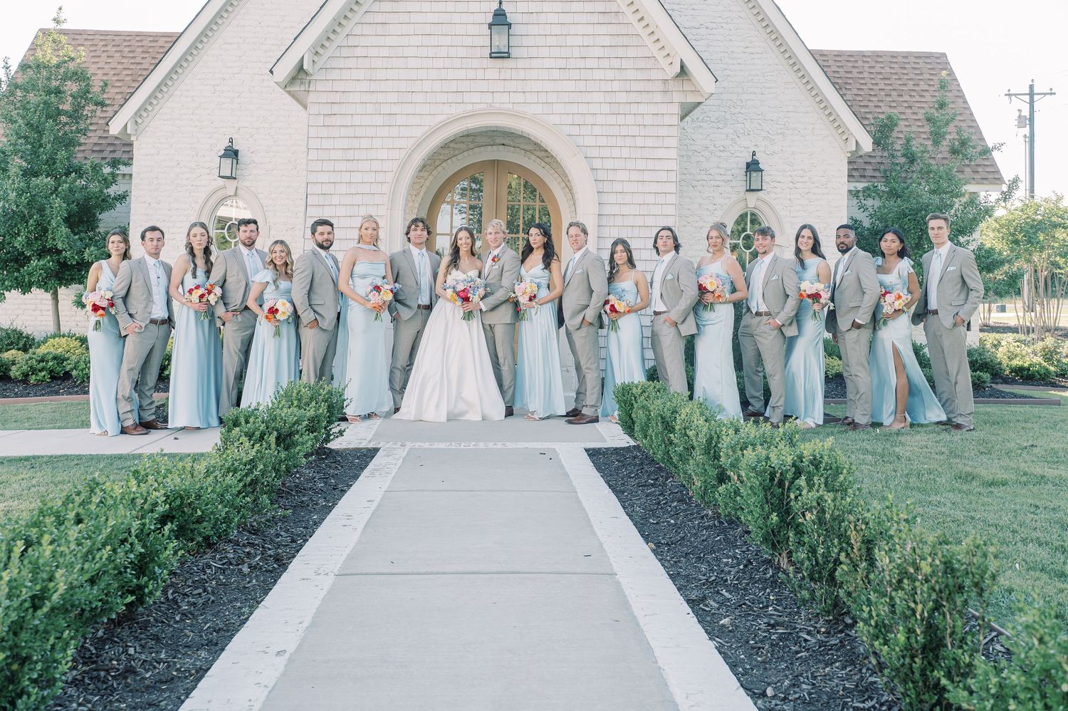 Wedding party poses in sage green and tan attire outside a white chapel with arched doorway and lush greenery.