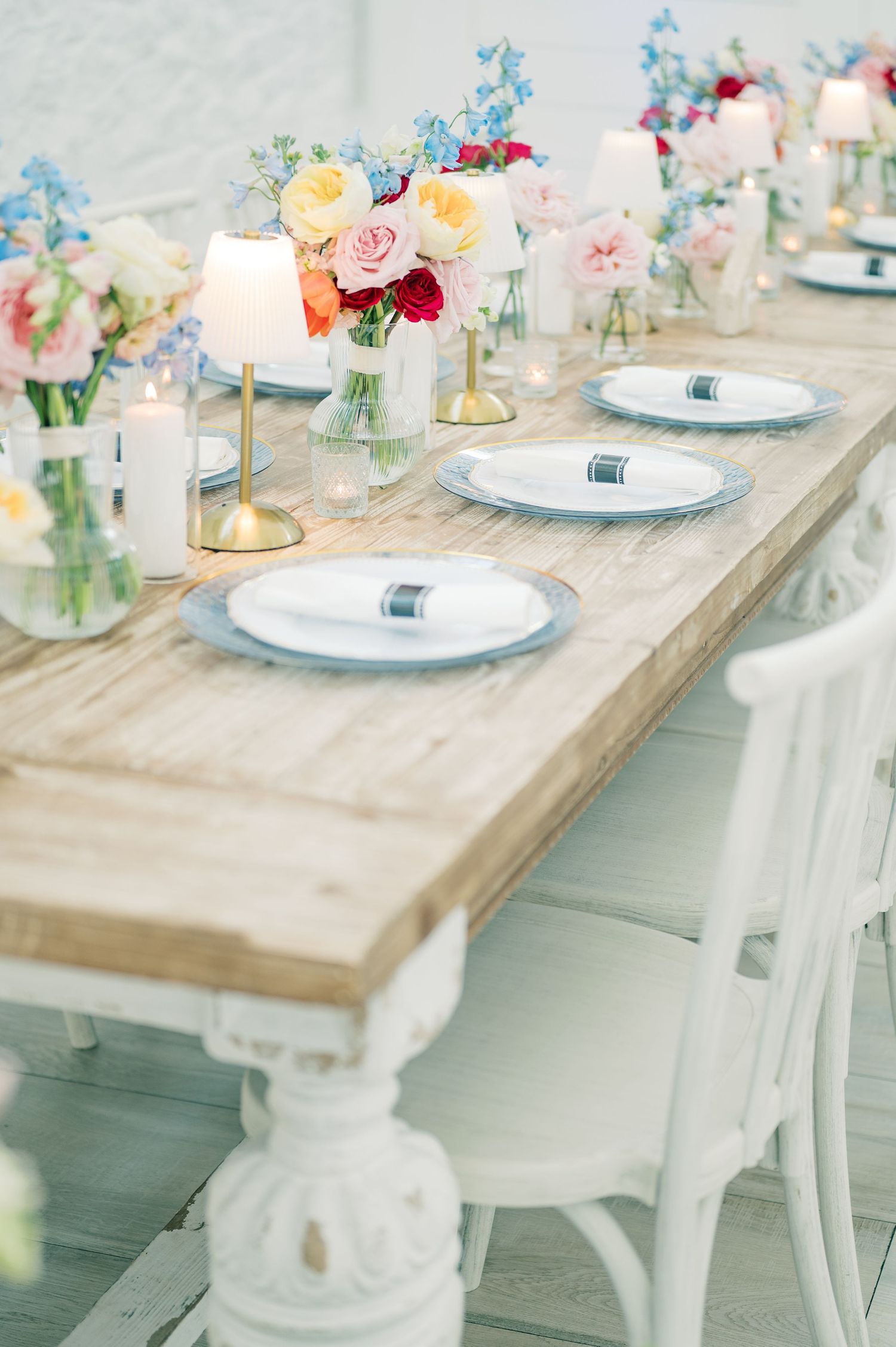 Elegant rustic wedding tablescape with colorful floral centerpieces, white charger plates, candles, and white chairs on a wooden farm table.